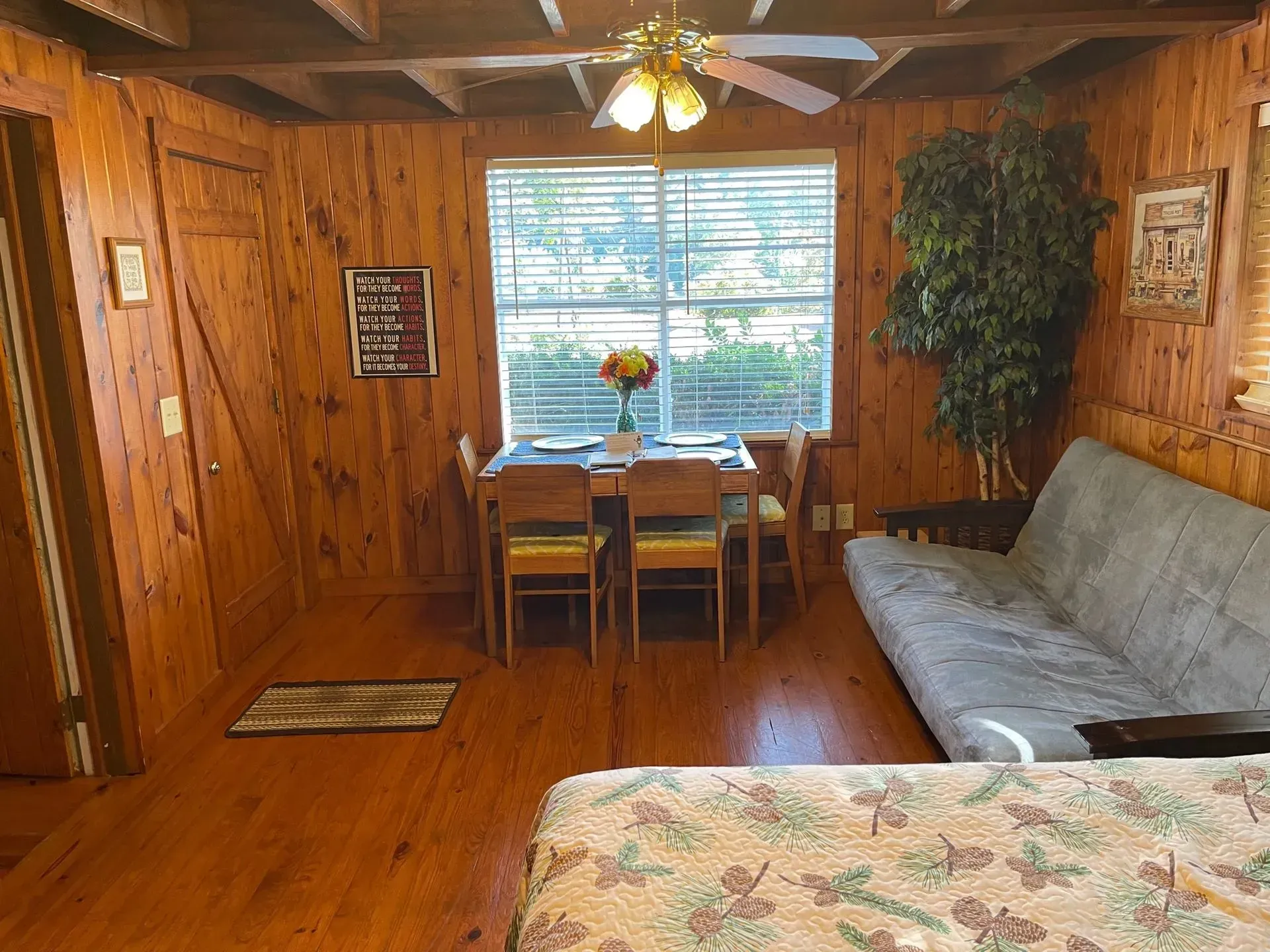 Cabin interior: wood-paneled walls, dining table with flowers, futon, small window, and a ceiling fan.
