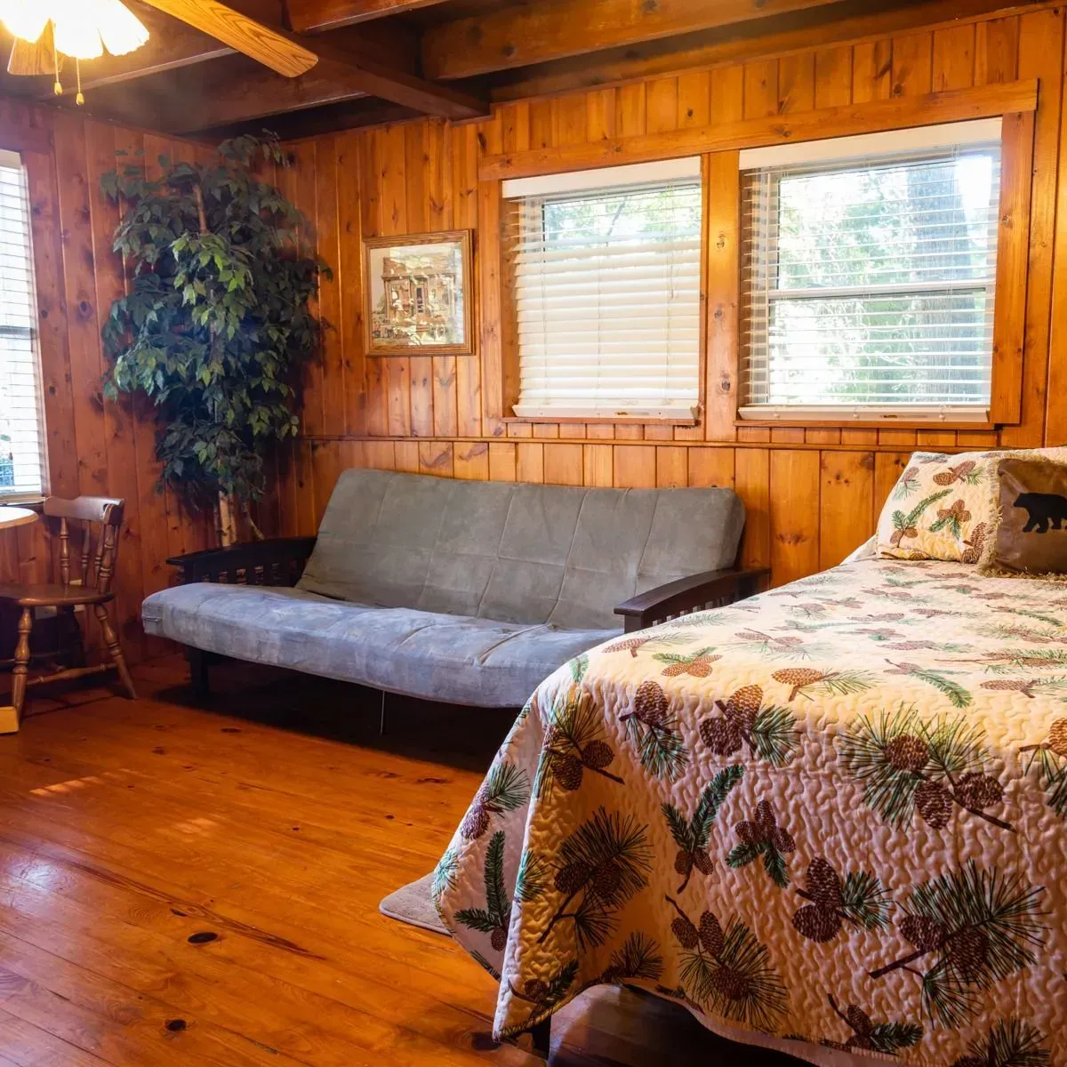 Cabin bedroom with wood paneling, bed, futon, windows, and a leafy tree.