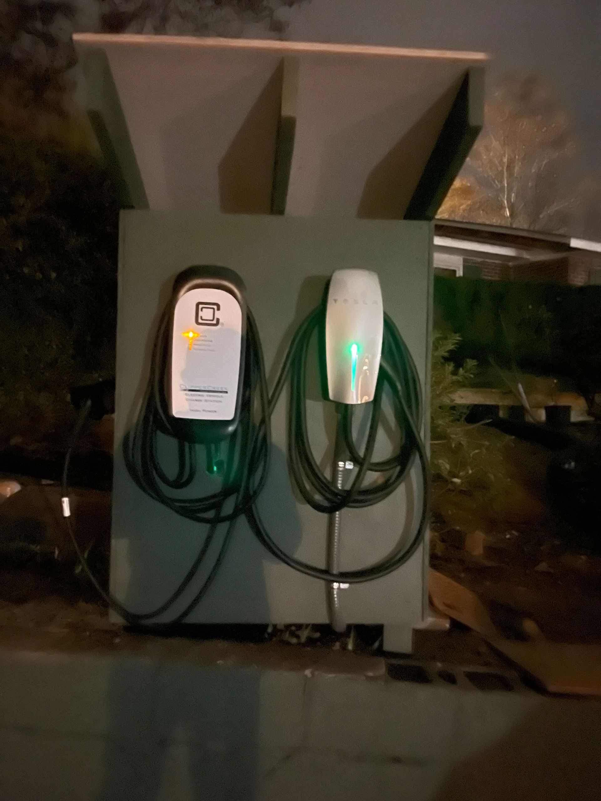 Two EV chargers mounted on a green box, cables coiled. One charger has a lit display, the other a green light.