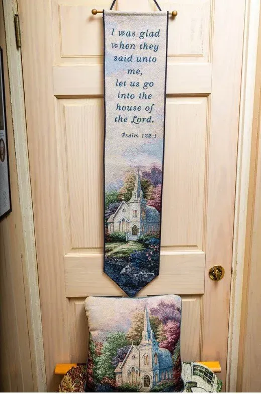 Banner with Bible verse over a church scene, above a pillow with the same scene, hung on a light wood door.
