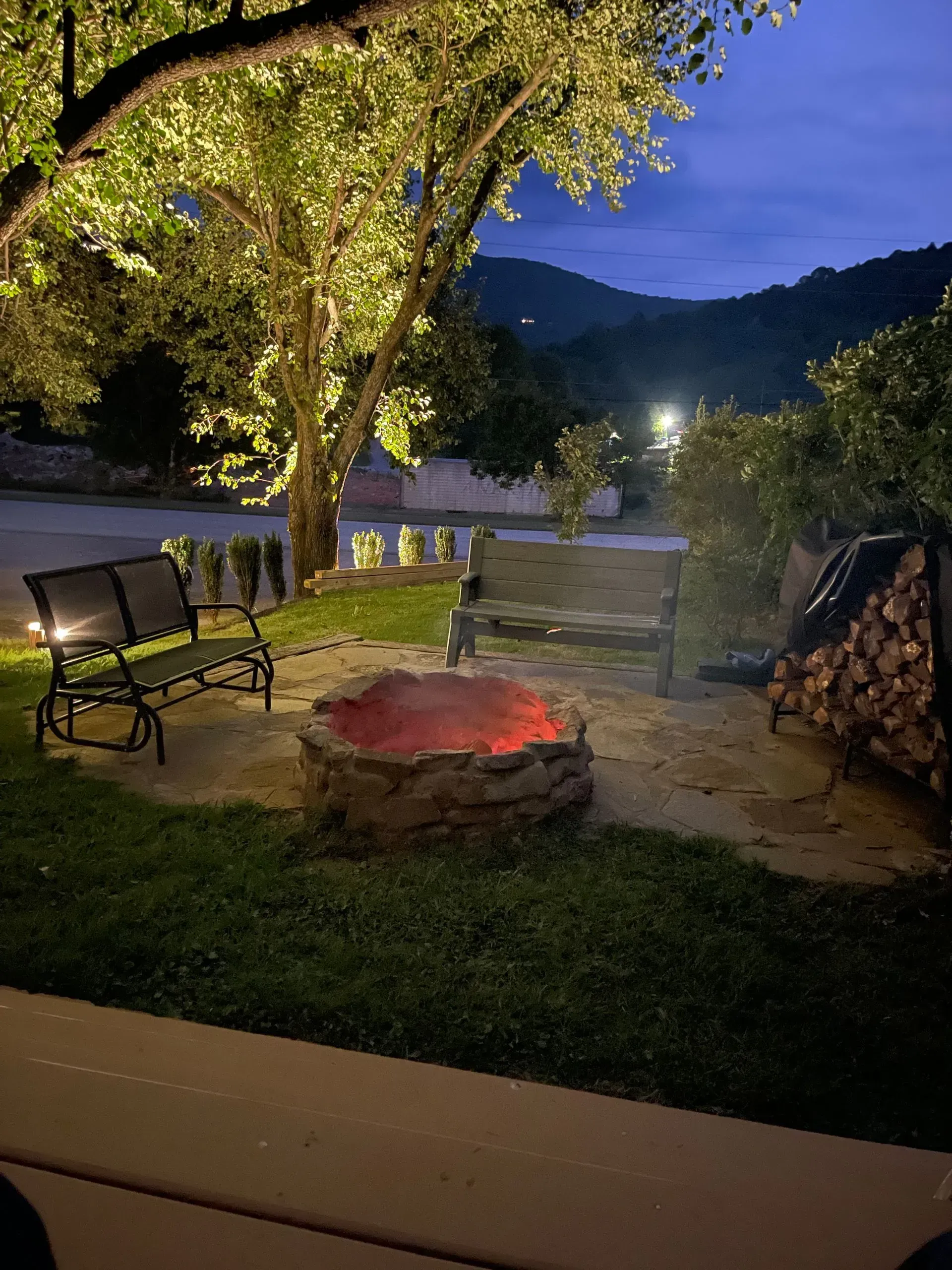 Fire pit with glowing embers surrounded by seating, lit tree, and a mountain backdrop.
