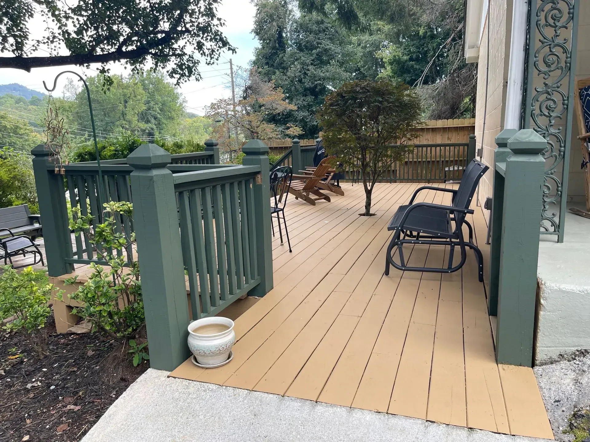 A wooden deck with green railings, tan floor. Black chairs and a small tree are on the deck.
