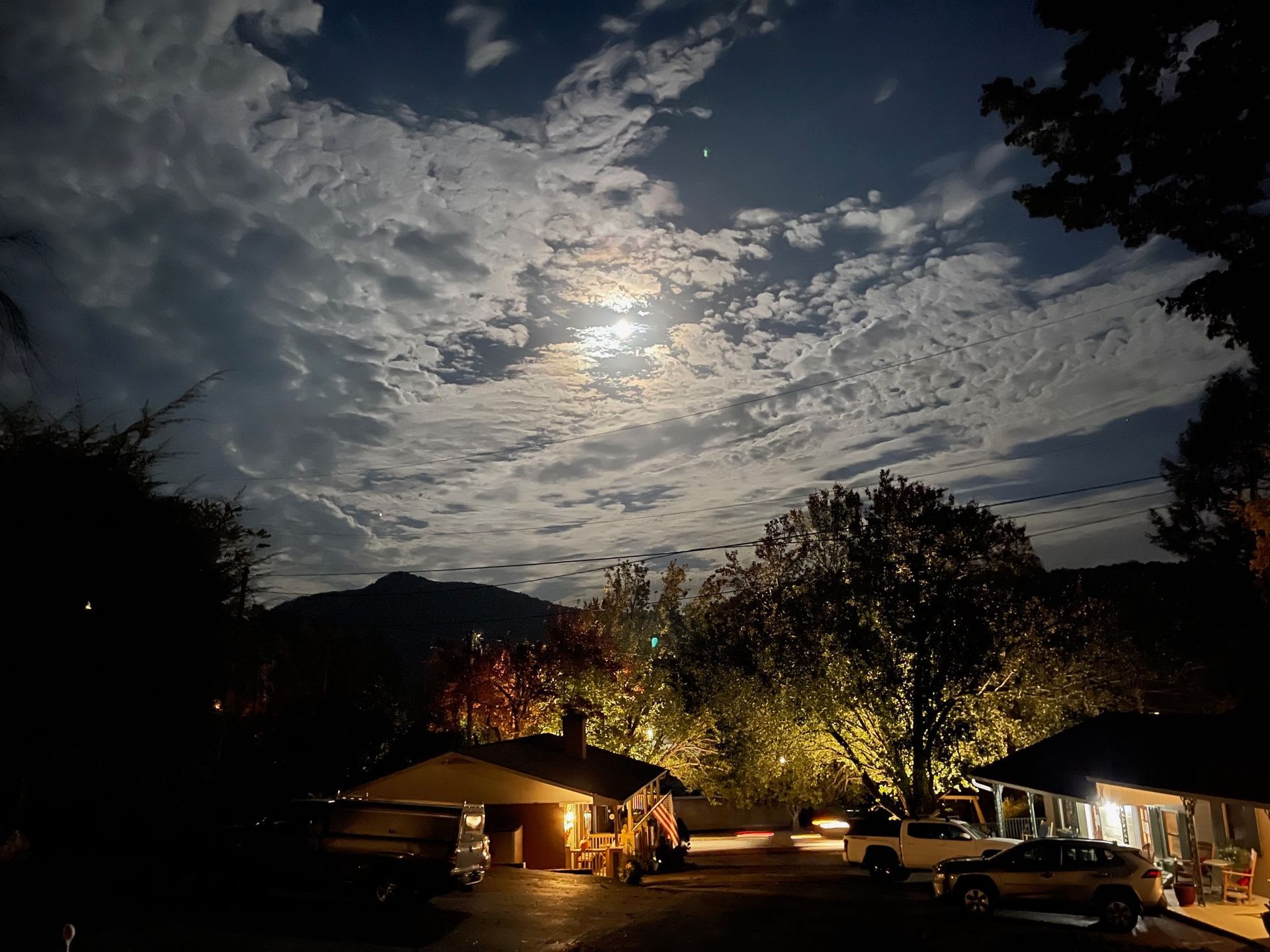 Night sky with full moon and clouds over houses and trees, lit by streetlights.