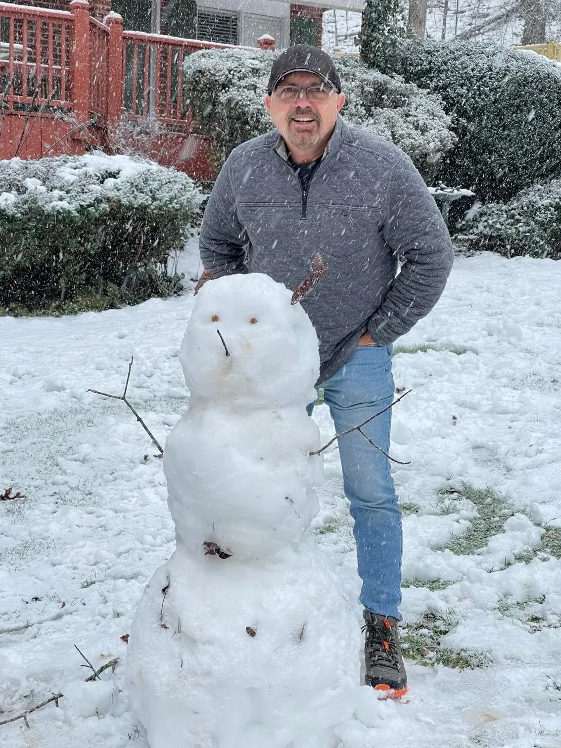 Man in gray sweater and jeans stands next to a snowman in the snow; trees and a house in the background.
