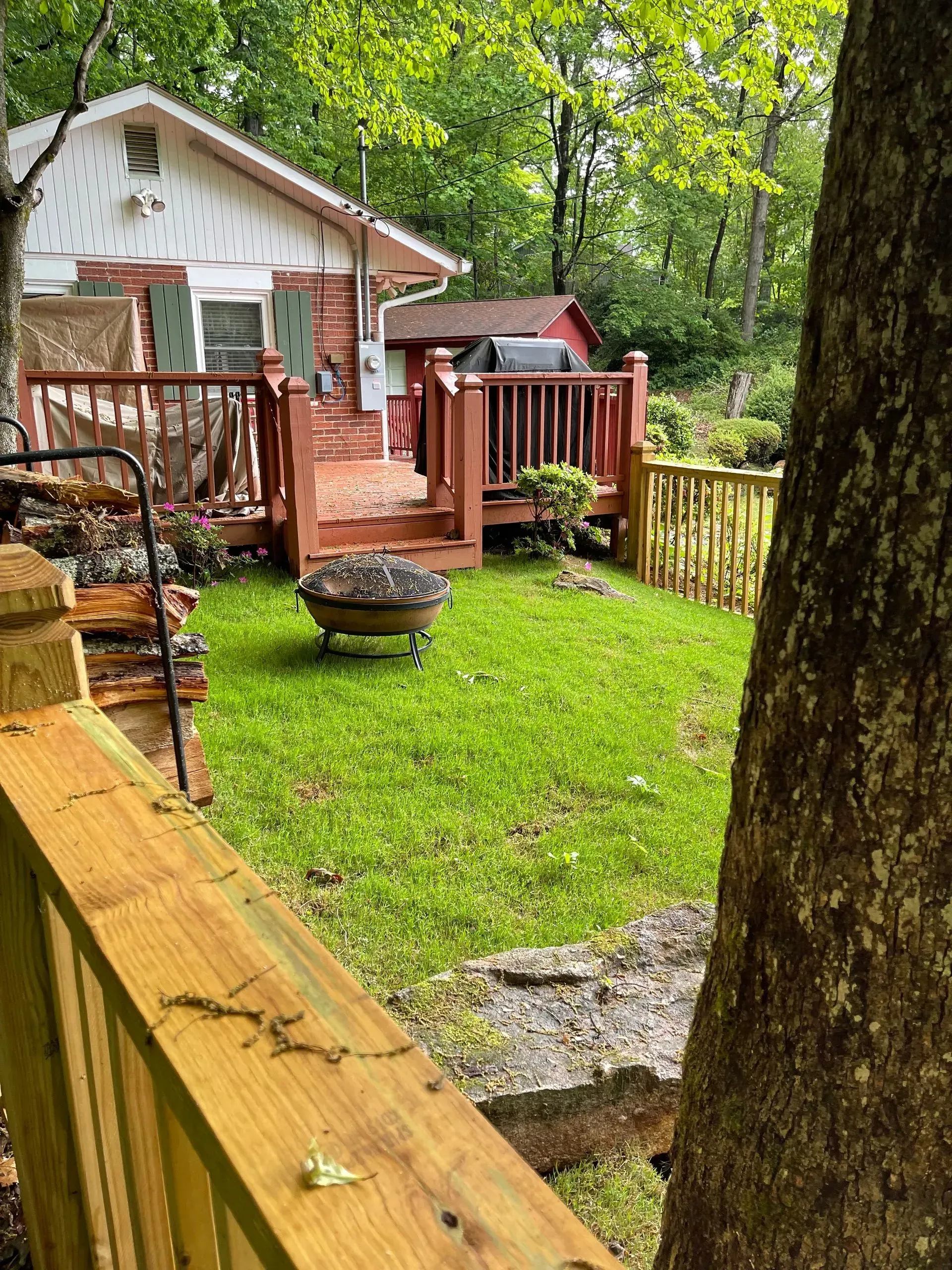 Small cottage with a wooden deck and a fire pit in a grassy backyard, surrounded by trees.