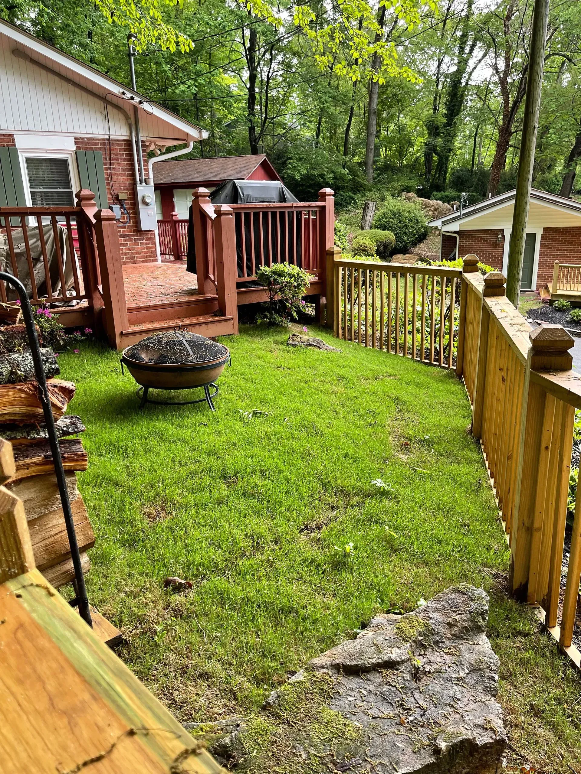 Lush green lawn with a fire pit, wooden deck, and fence, near a house in a wooded area.