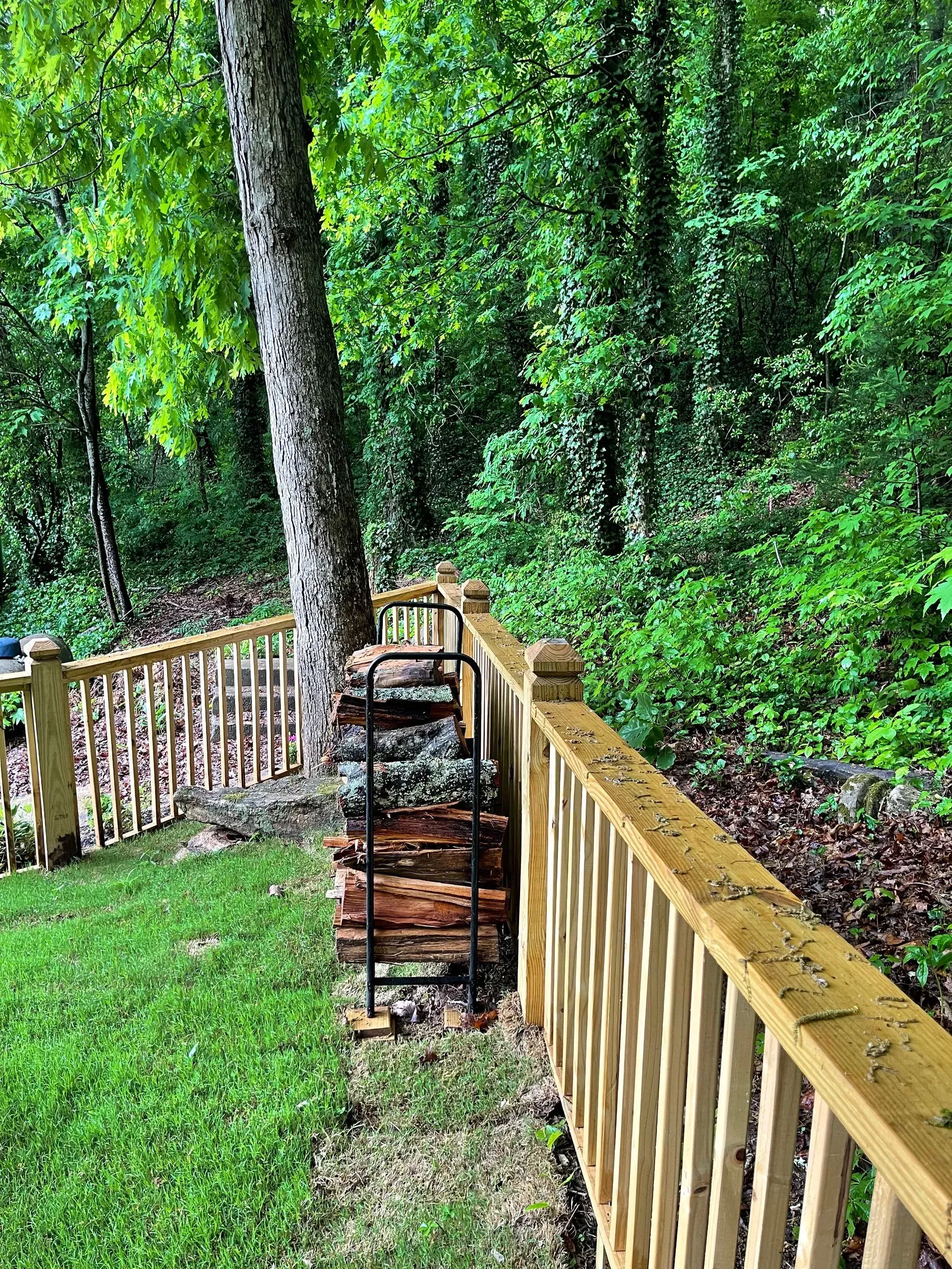 Green backyard with a woodpile against a fence. A tree and wooded hill are in the background.