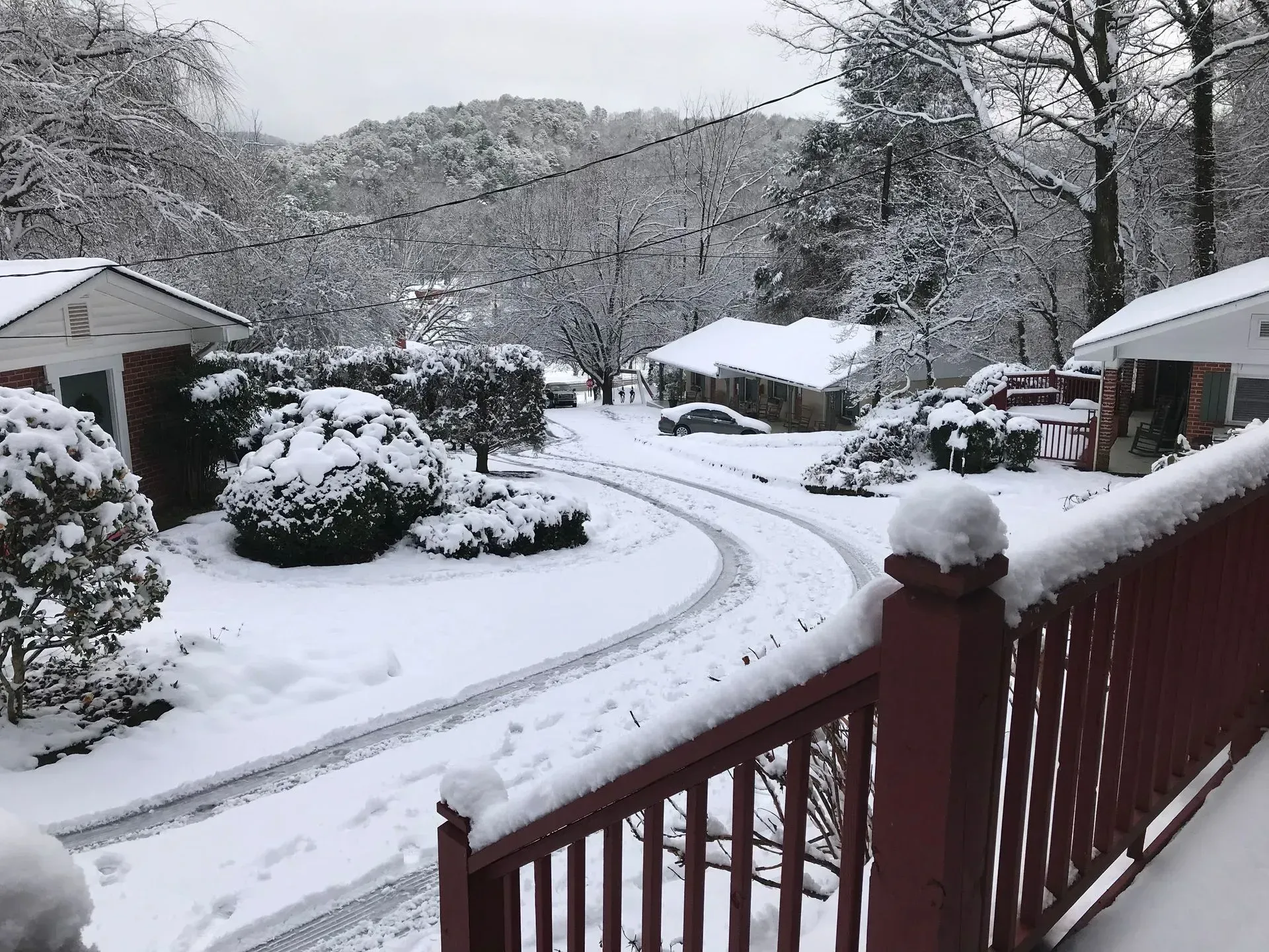 Snowy residential area, curved driveway, red porch railing, white houses, snow-covered trees and bushes, mountain background.