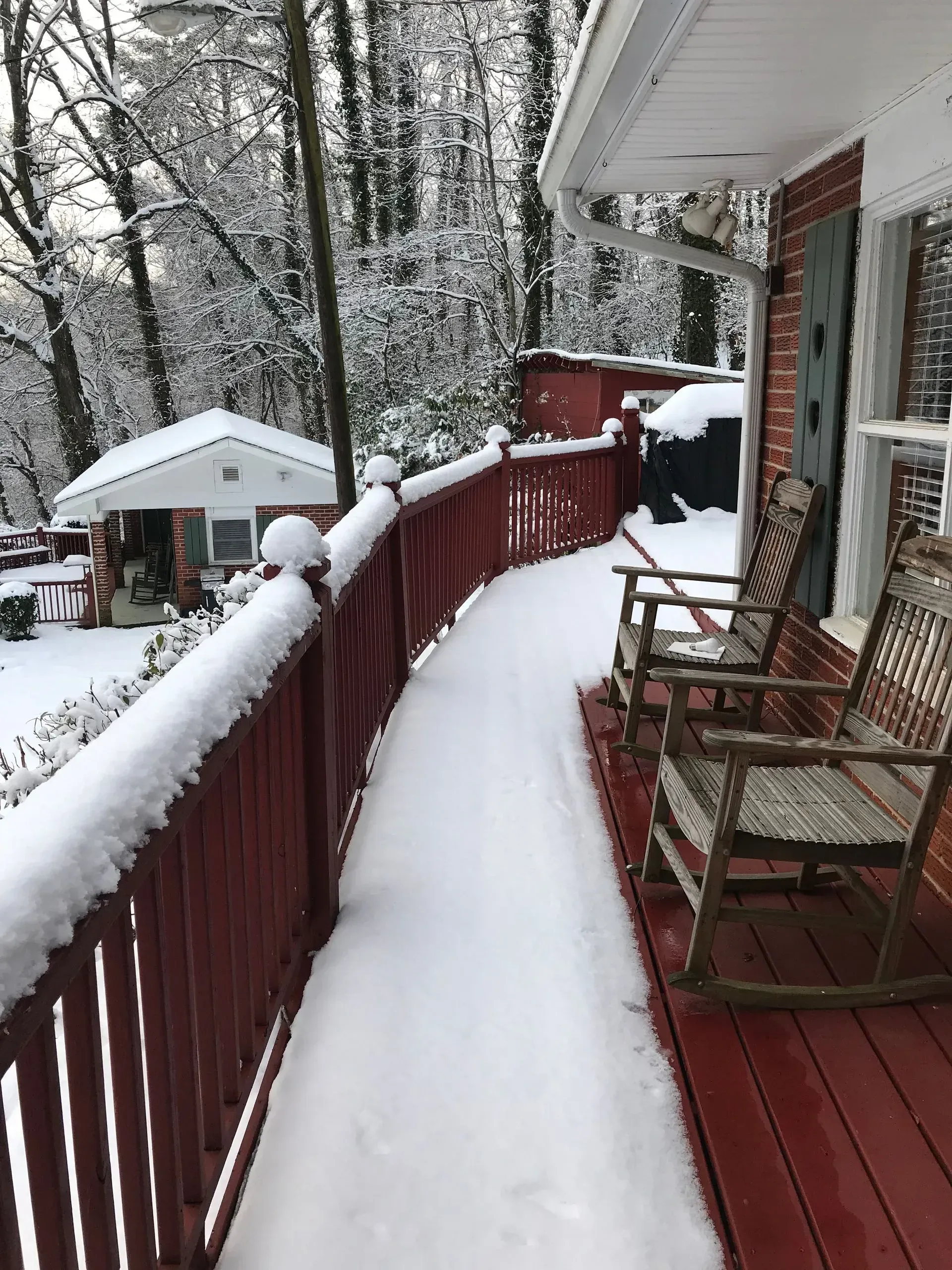 Snow-covered porch with red railing, two rocking chairs, and a small building in a wintery forest setting.