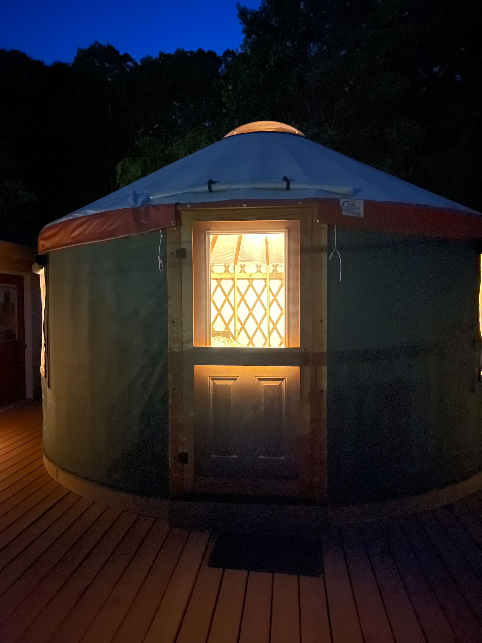 Yurt at dusk with lit window and door, orange trim, on a wooden deck.
