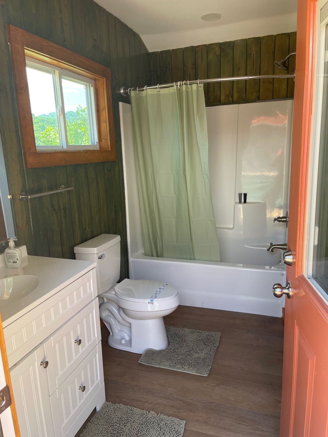 Small bathroom with green walls, white tub/toilet, wooden floor, and orange door.