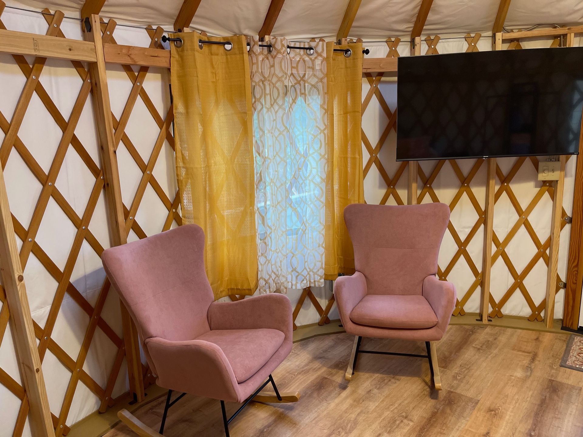 Two pink armchairs facing a window with gold curtains in a yurt. TV on the wall.