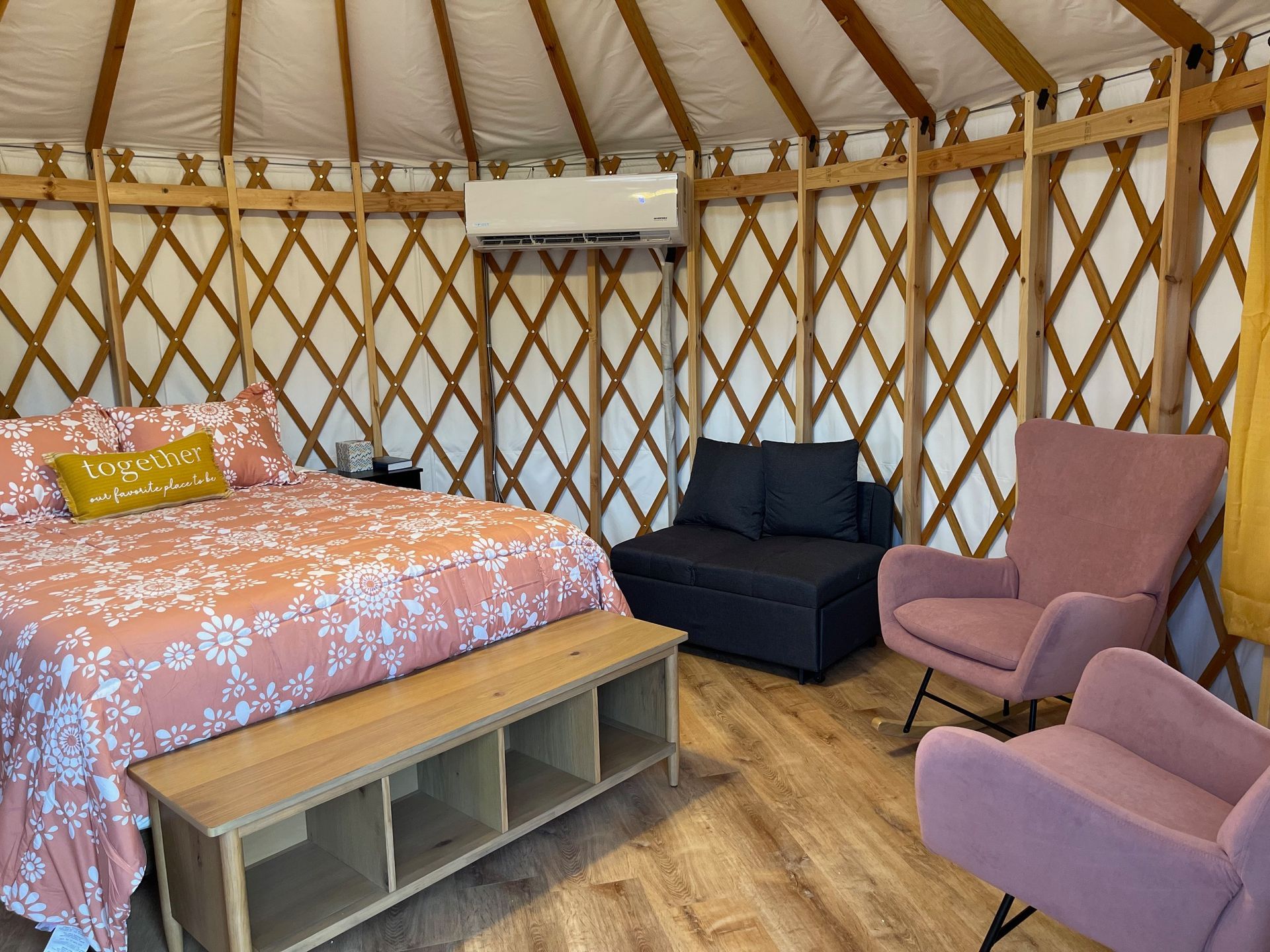 Interior of a yurt with bed, seating area, and air conditioning. Soft pink and wood tones.