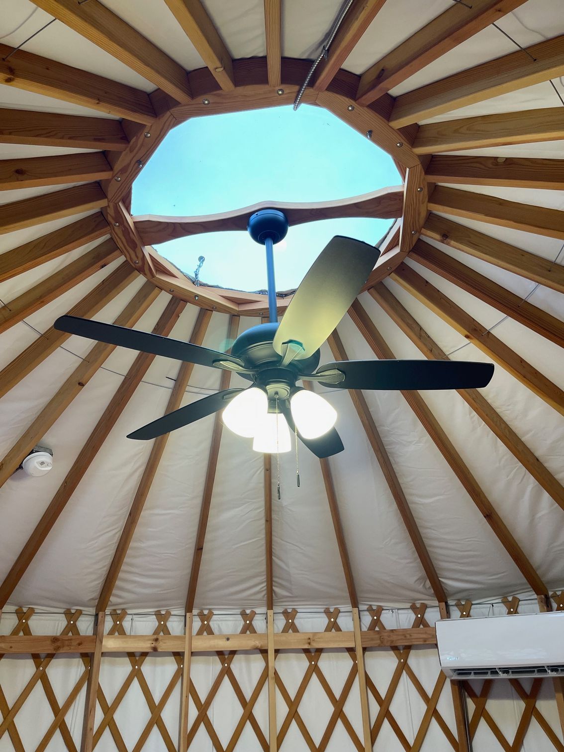 Ceiling fan in a yurt with a central skylight. Wooden beams radiate from the opening.