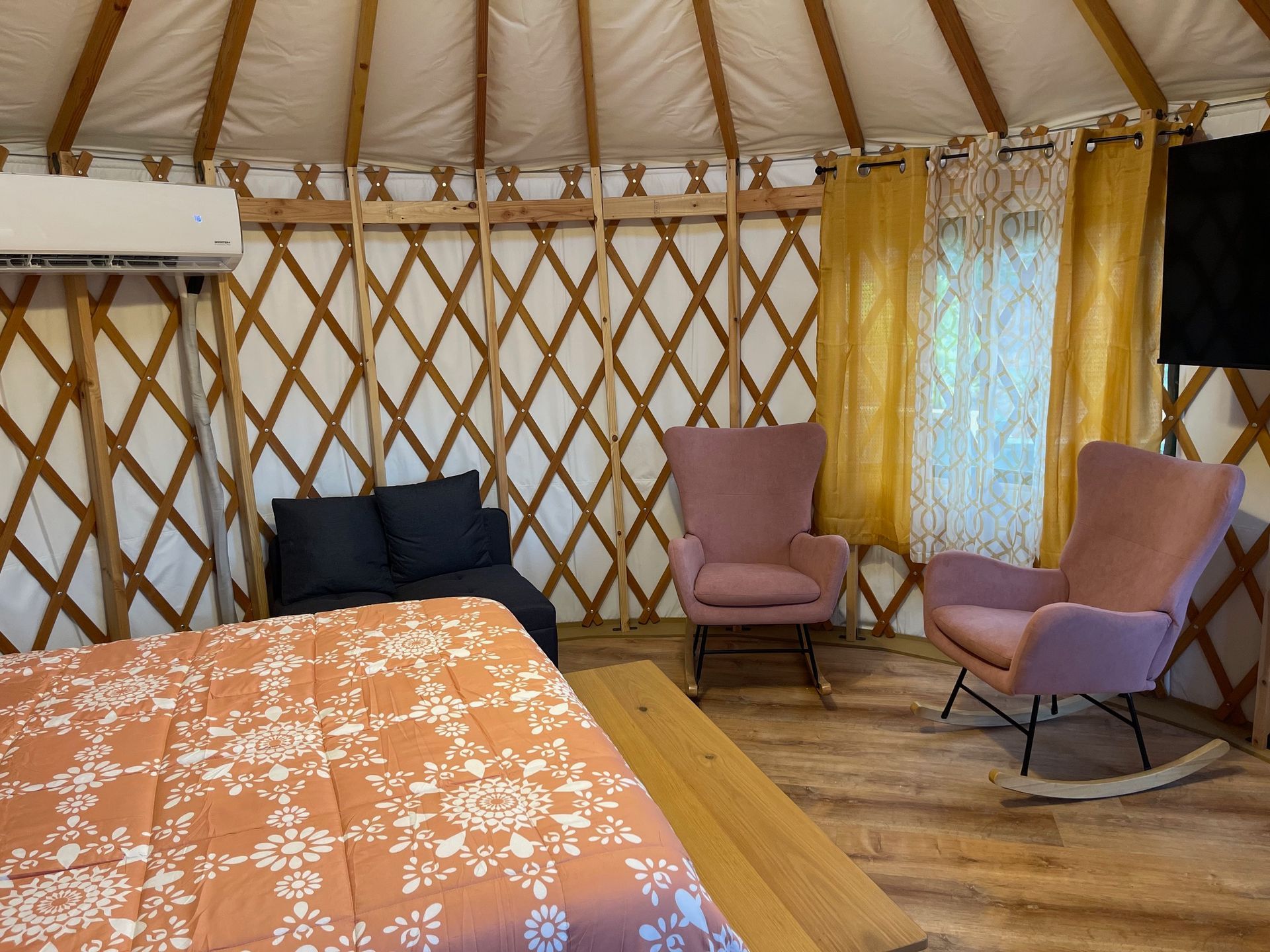 Interior of a yurt with a bed, seating area, and air conditioning unit. Warm colors and natural light.