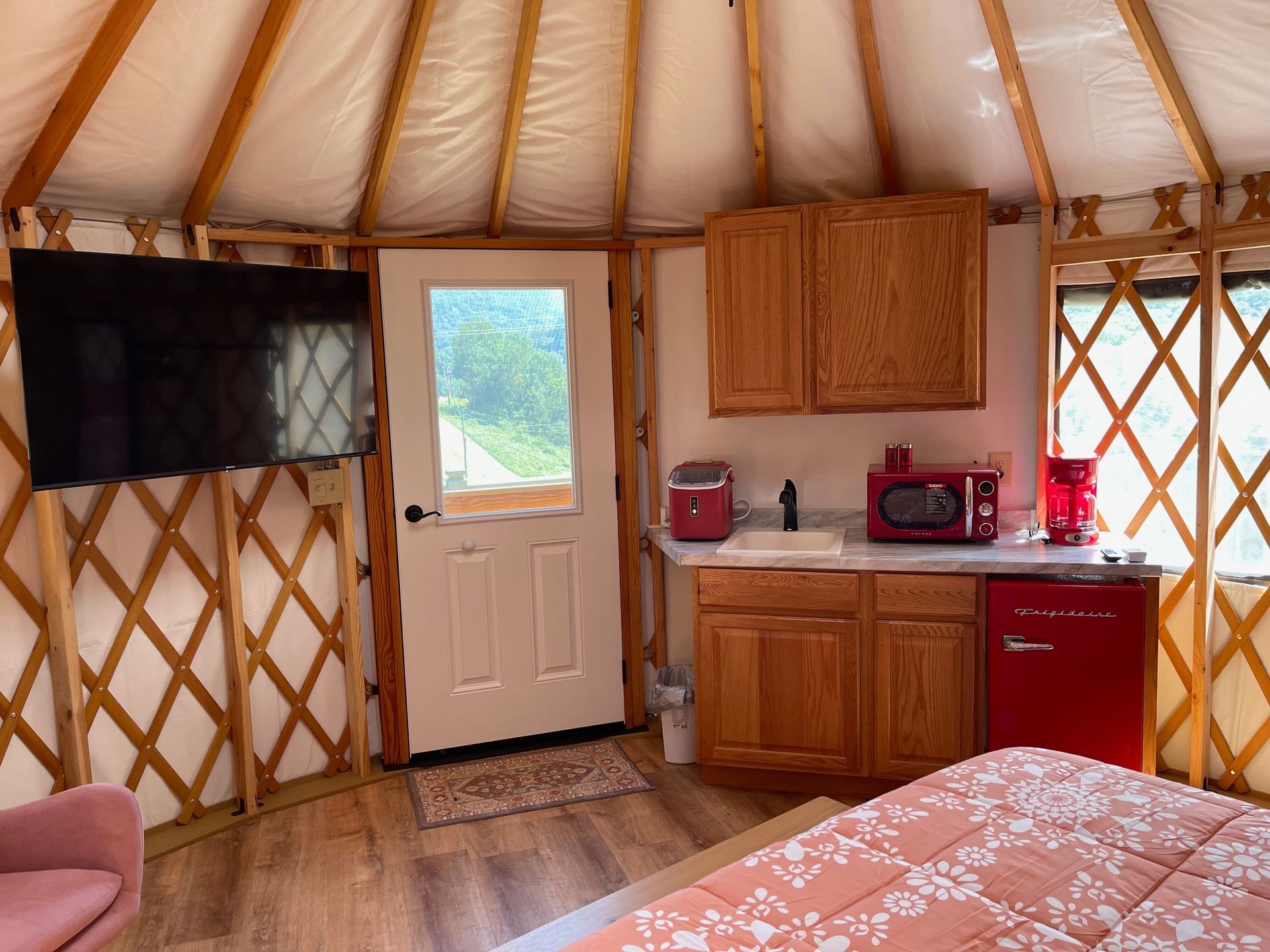 Interior of yurt with TV, kitchenette, and bed. Wooden beams, light brown cabinets, and red appliances.