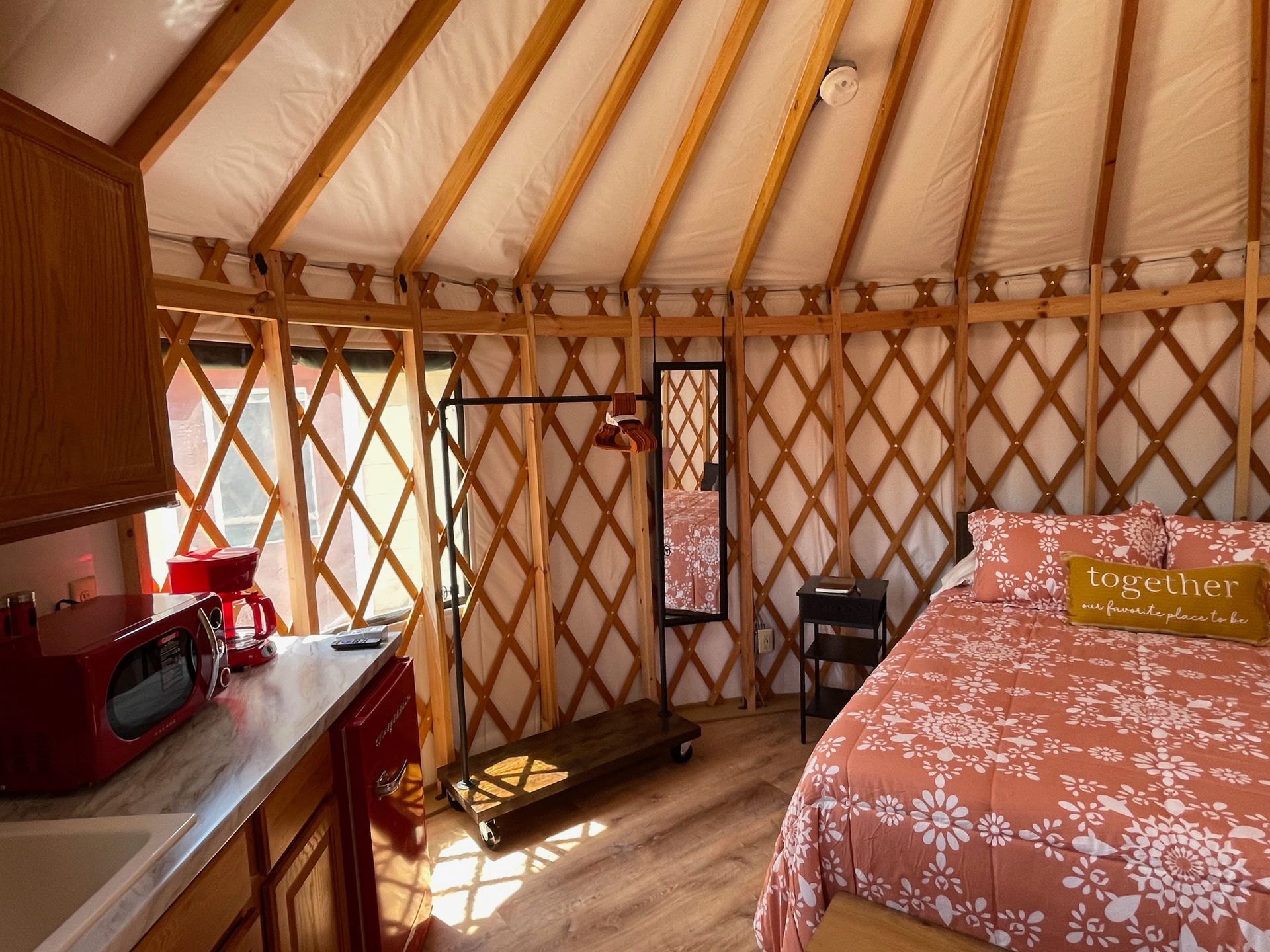 Interior of a yurt: bed, kitchen, mirror, clothing rack, wood lattice walls, and natural light.
