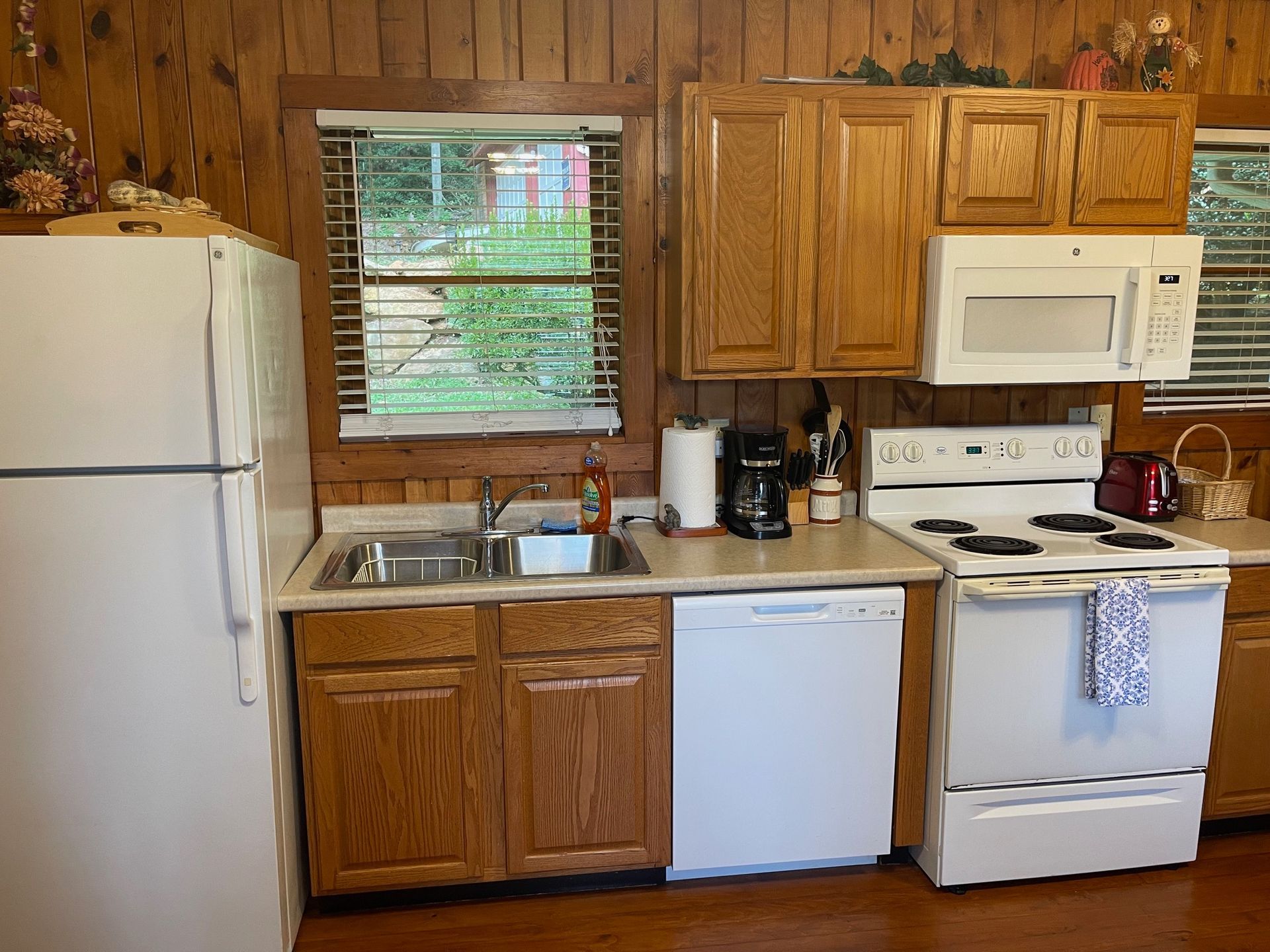 Small kitchen with wood paneling, appliances (fridge, stove, microwave), cabinets, windows, and a TV.