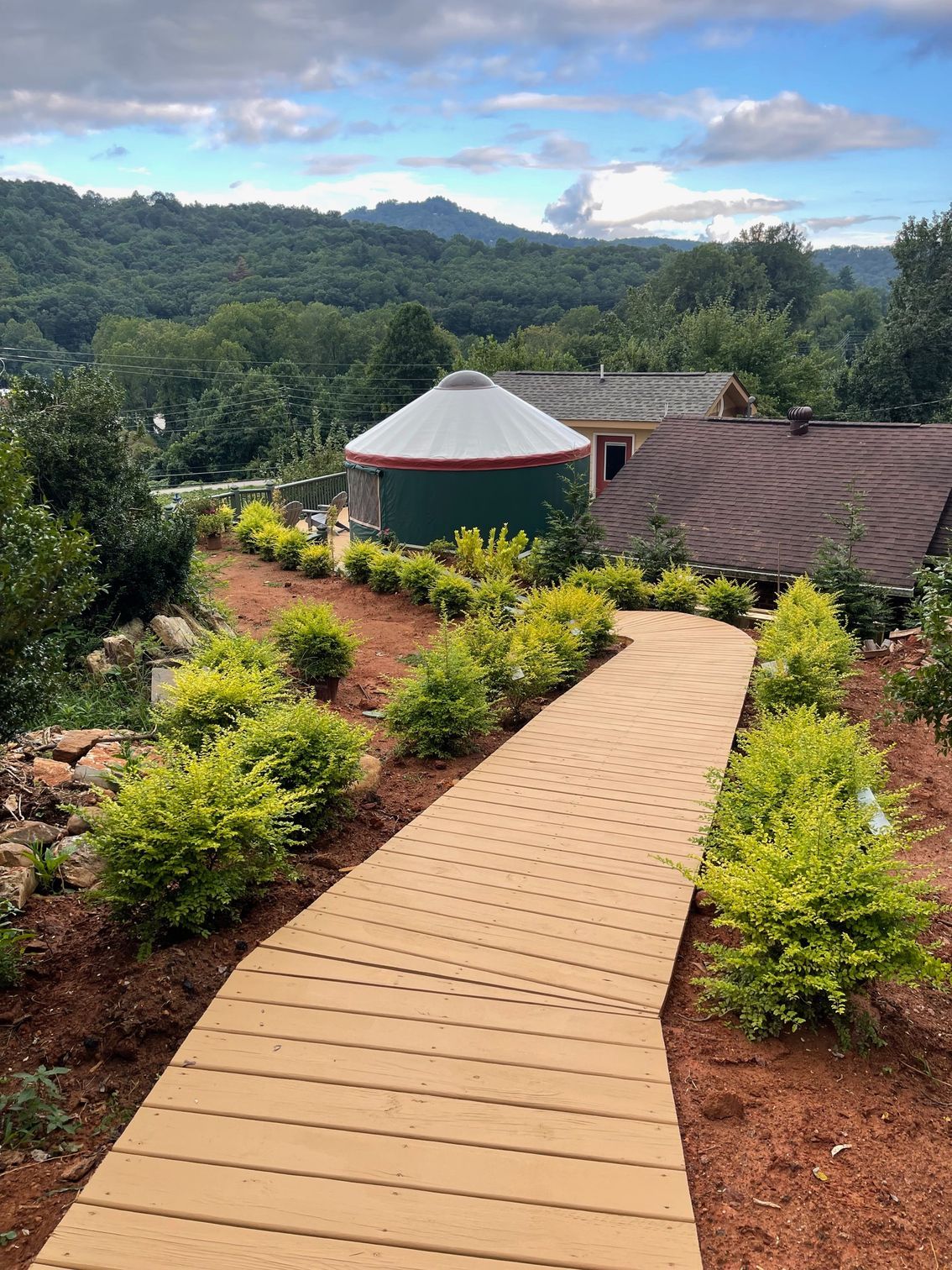 Wooden walkway leads to yurt with roof extension; lush green foliage surrounds. Mountain background, blue sky.