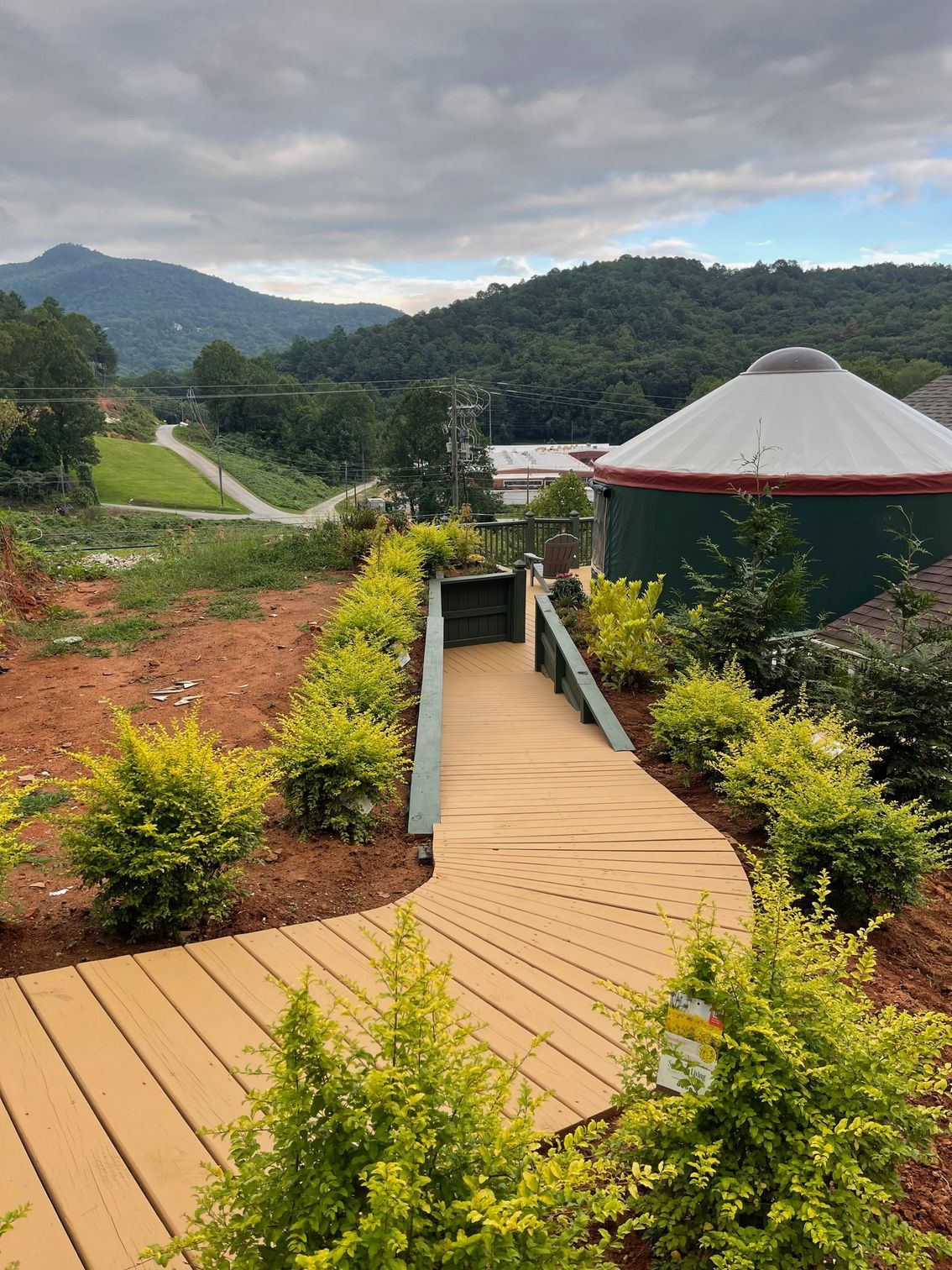 A yurt and house sit on a hillside with a wooden walkway. A black cat is in the foreground.