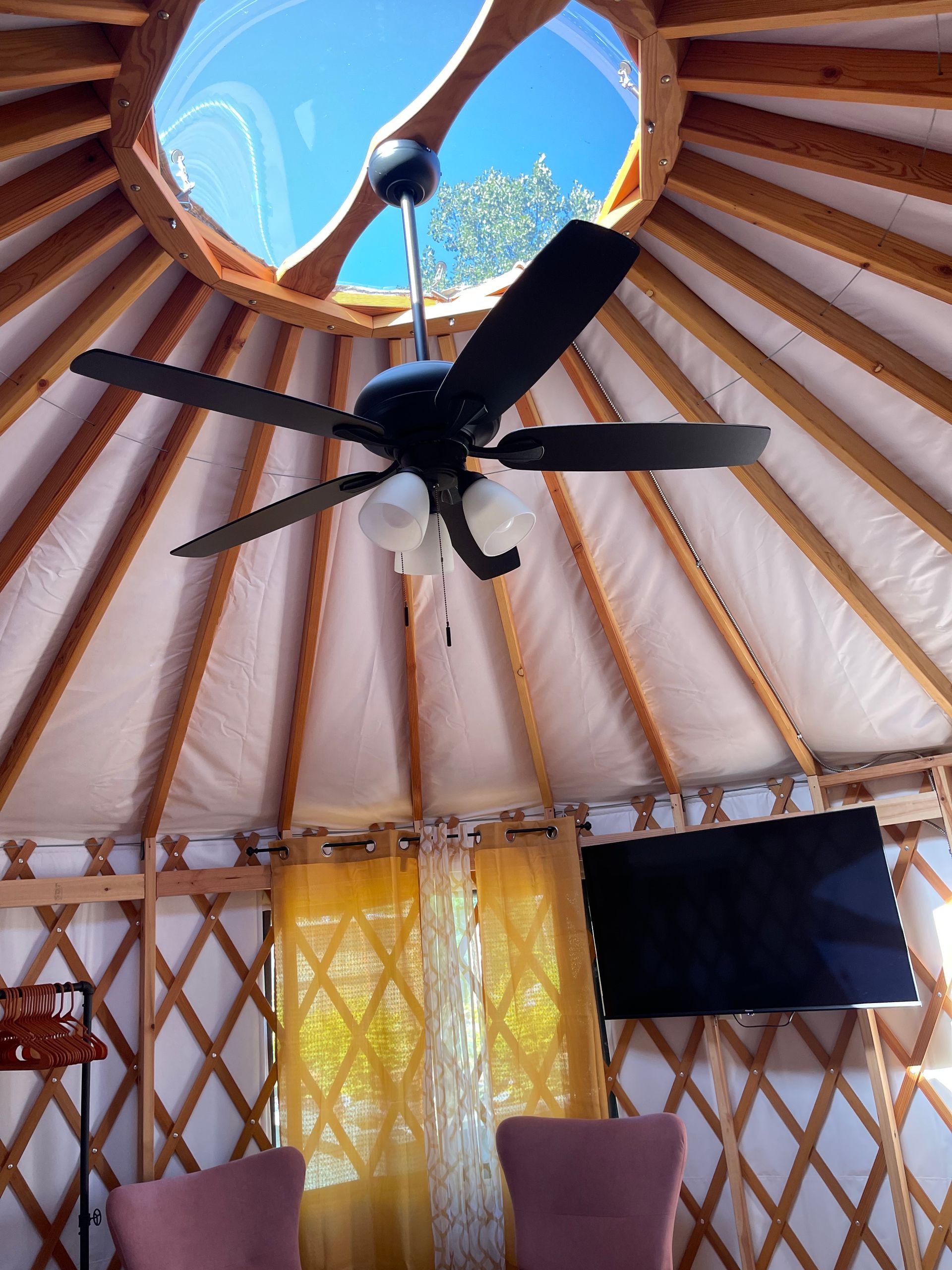 Inside of a yurt. A ceiling fan, skylight, and TV. Wooden lattice walls with pink chairs.