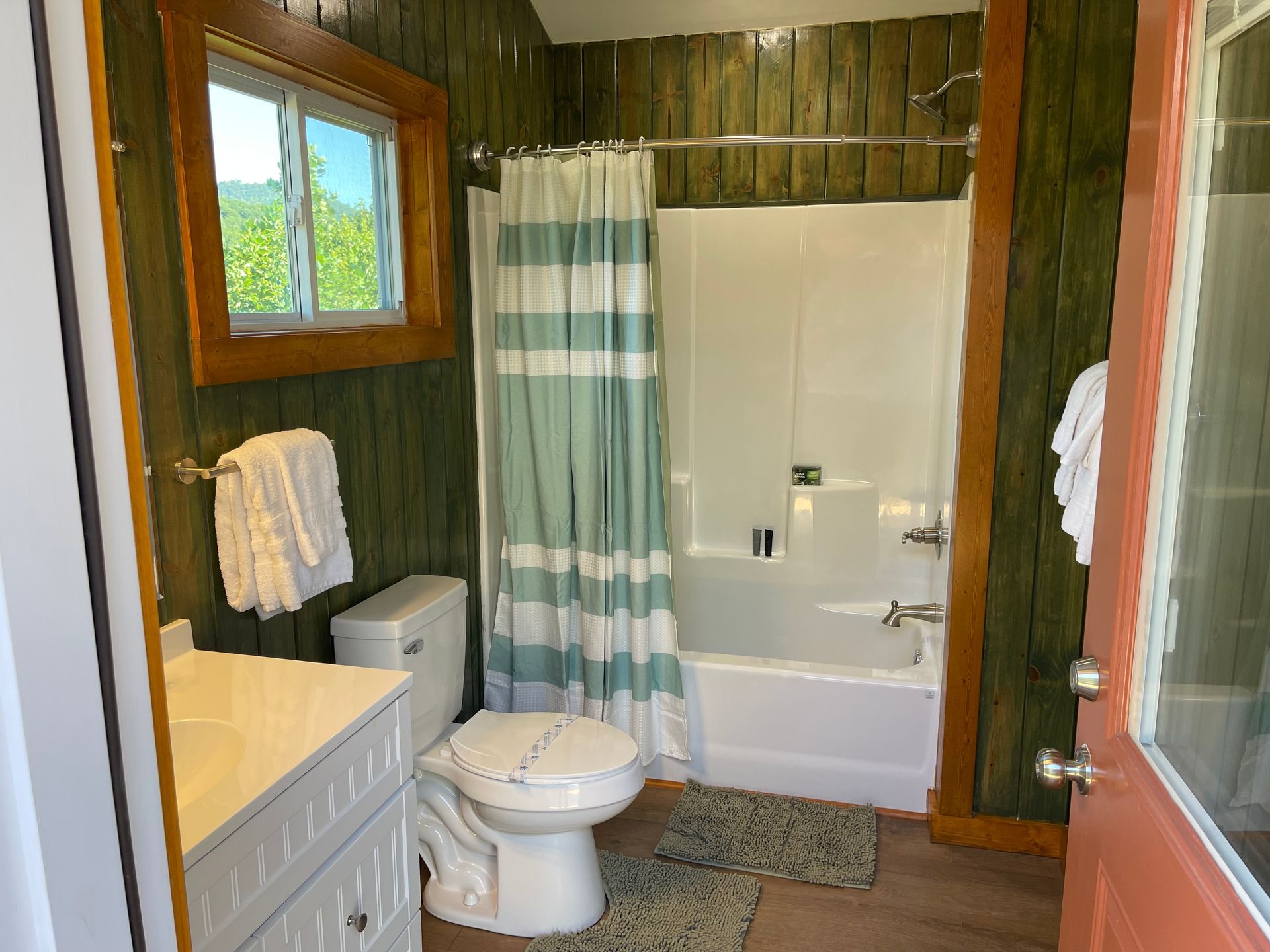 Small bathroom with green wood paneling, a white tub and toilet, and a window.