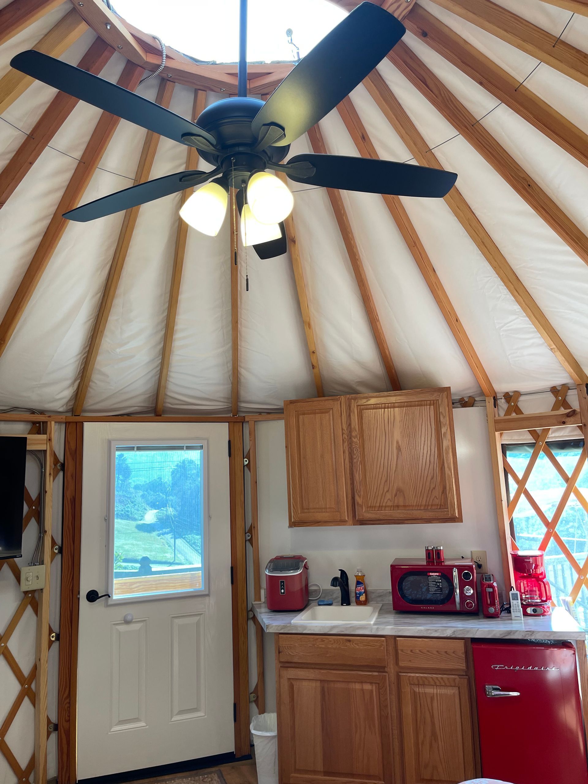 Interior of a yurt with a kitchen. Includes ceiling fan, cabinets, red appliances, and a door to the outdoors.