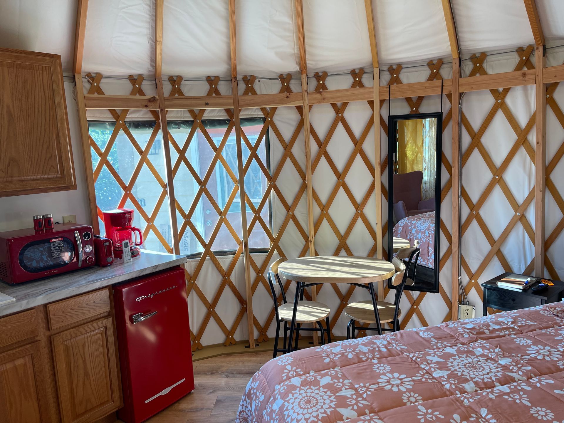 Interior of a yurt with bed, table, kitchenette, and mirror. Red accents. Wooden lattice walls.