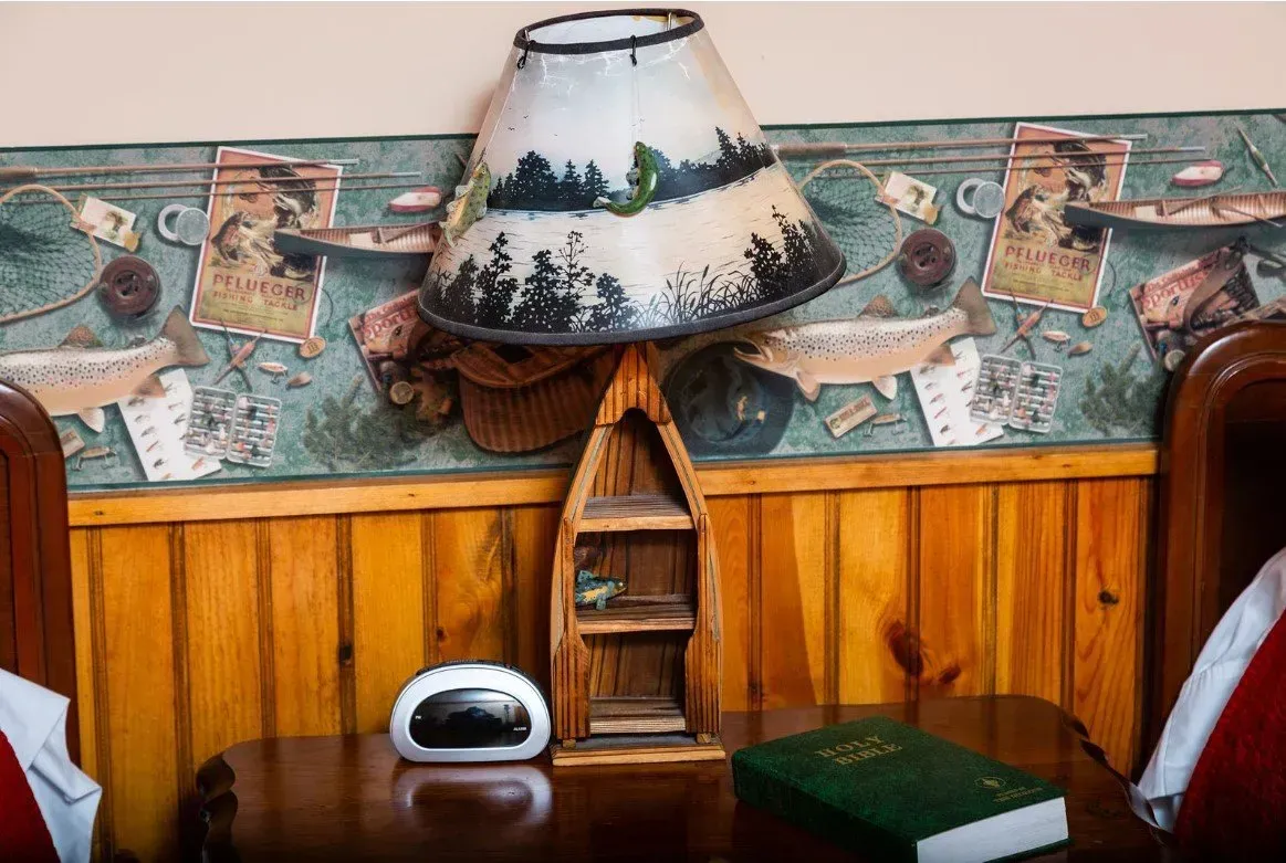 A wooden nightstand with a lamp, alarm clock, and book against a wood-paneled wall with rustic wallpaper.