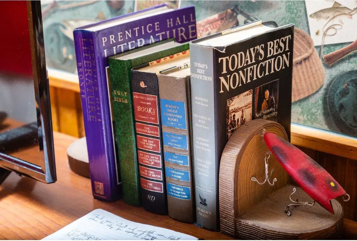 Books of various colors stand on a wooden desk, next to a fishing lure bookend.