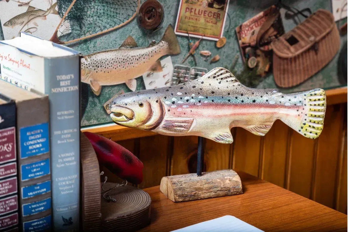 Wooden fish carving on a table next to books and a fishing vest.