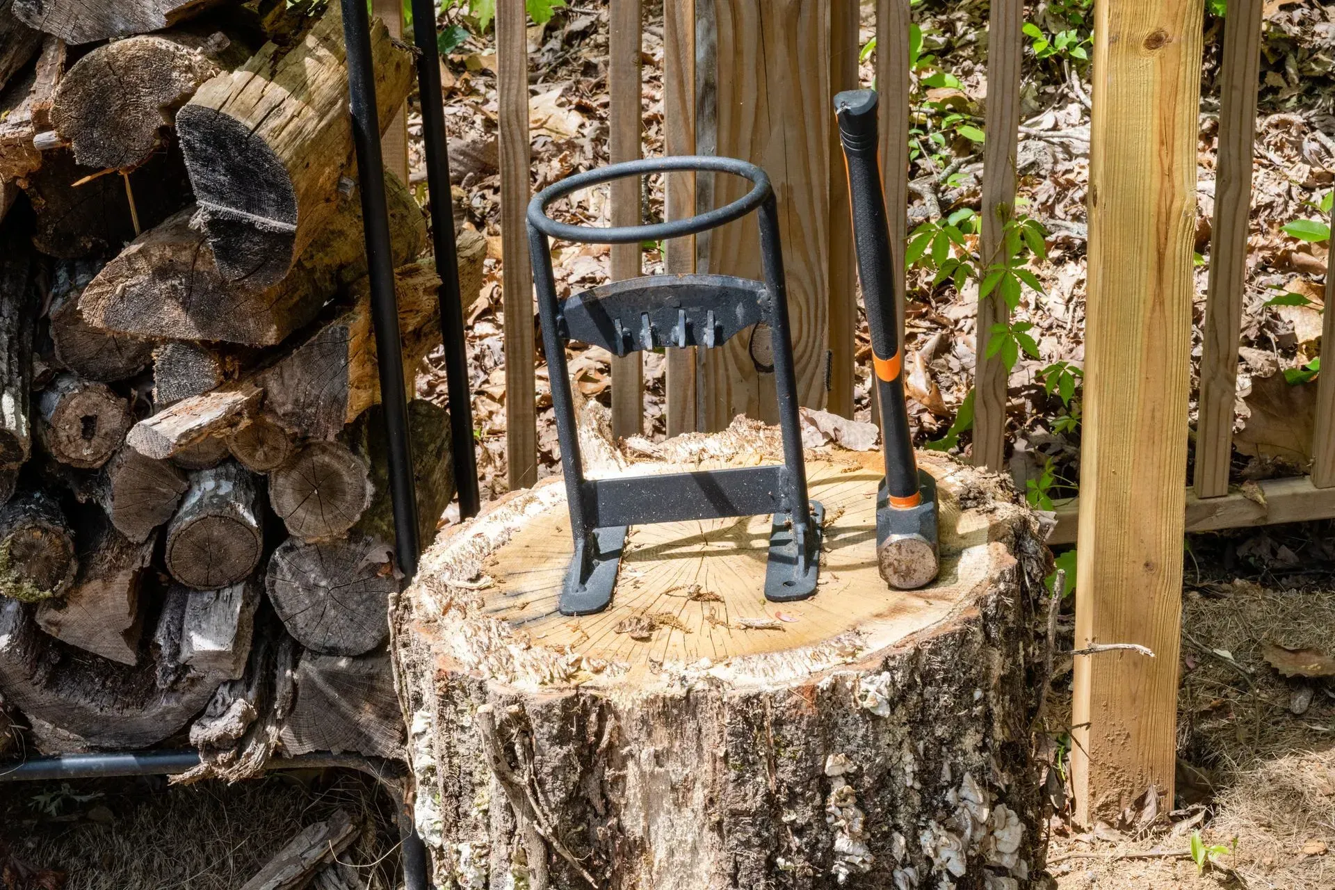 Log-splitting tools on a tree stump: metal splitter, sledgehammer, and stacked firewood in a backyard.