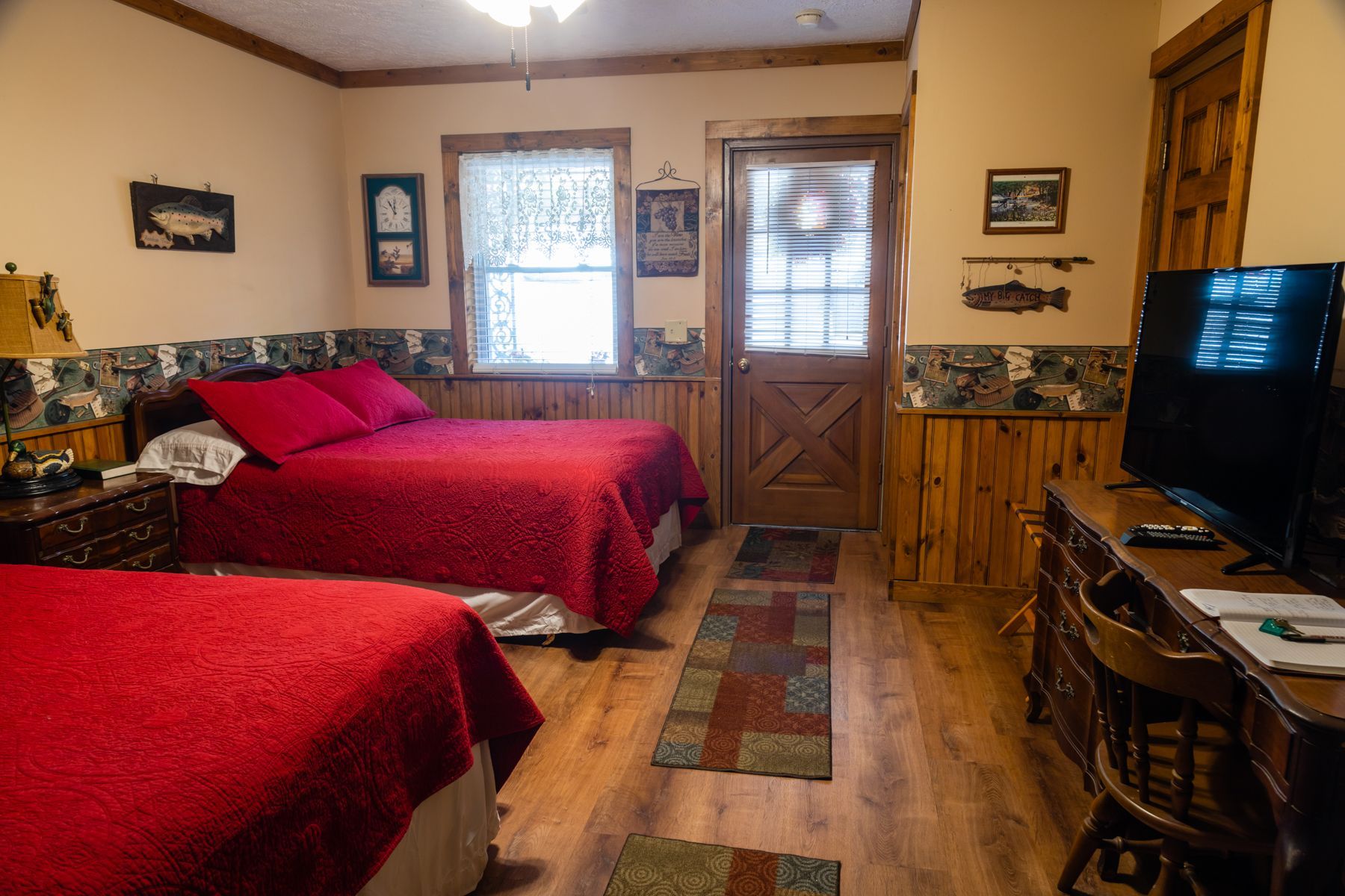 A rustic bedroom with two beds, a TV, and a wooden door. Red bedspreads, wood paneling.