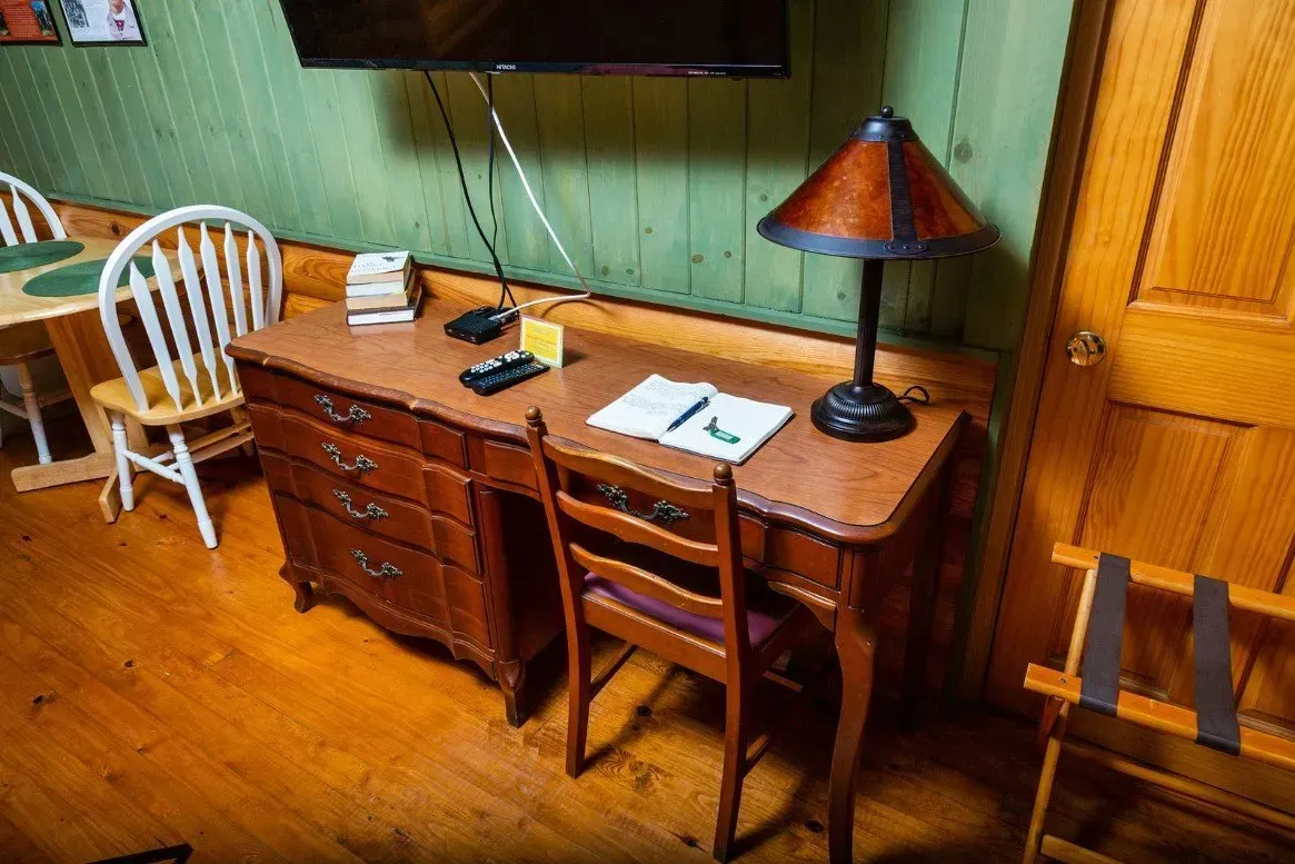 Wooden desk with drawers and chair in a room with a small table and lamp; luggage rack beside a door.