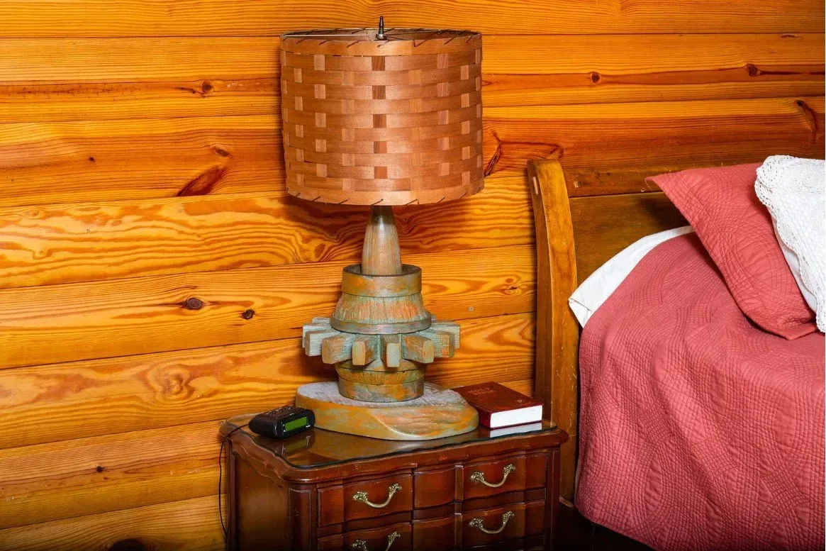 Lamp with woven shade on a wood nightstand next to a bed with a red quilt, in a room with wood paneling.