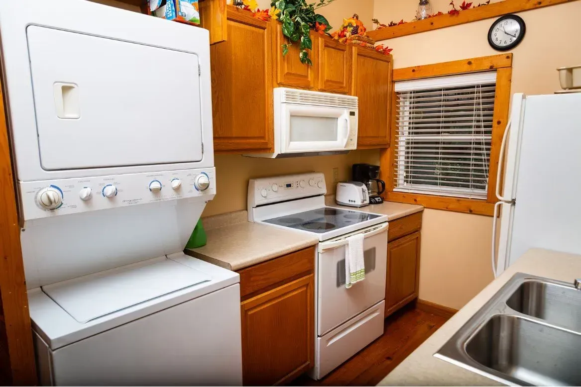 Cozy kitchen with appliances: stacked washer/dryer, oven, microwave, toaster, and sink. Brown cabinets, cream countertops.