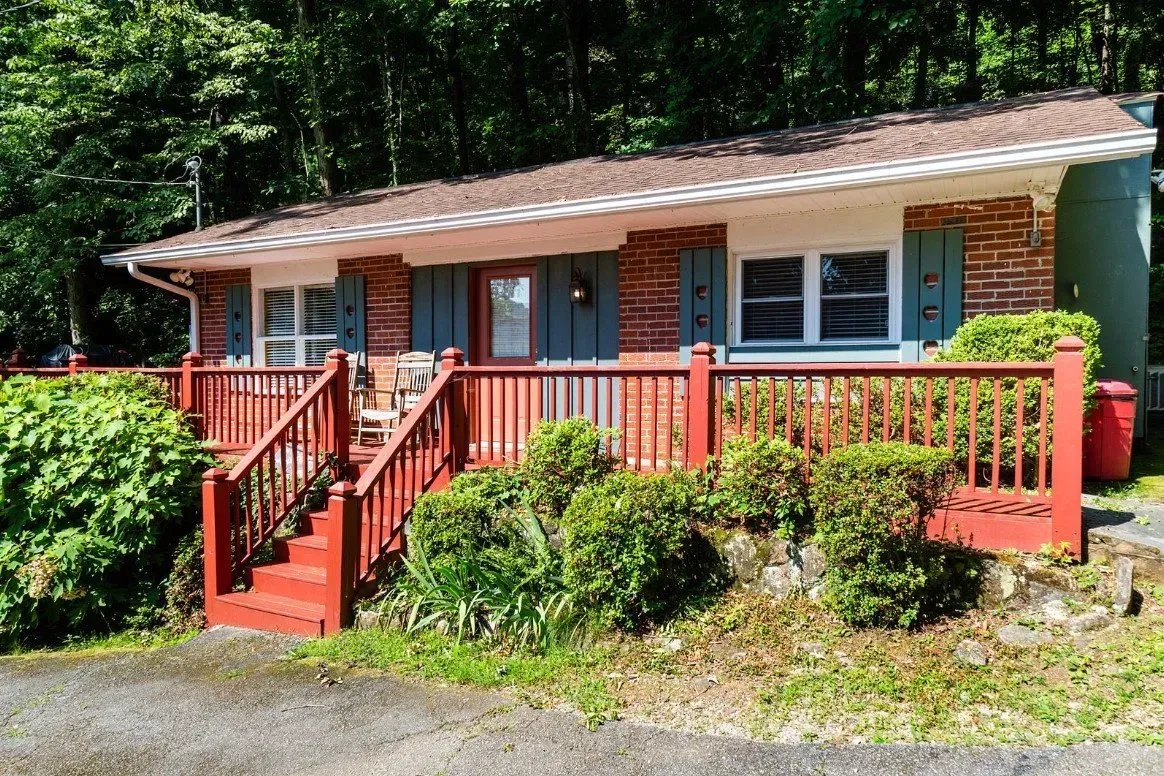 Red-trimmed cottage with brick facade and green shutters, front porch with railing, surrounded by greenery and trees.