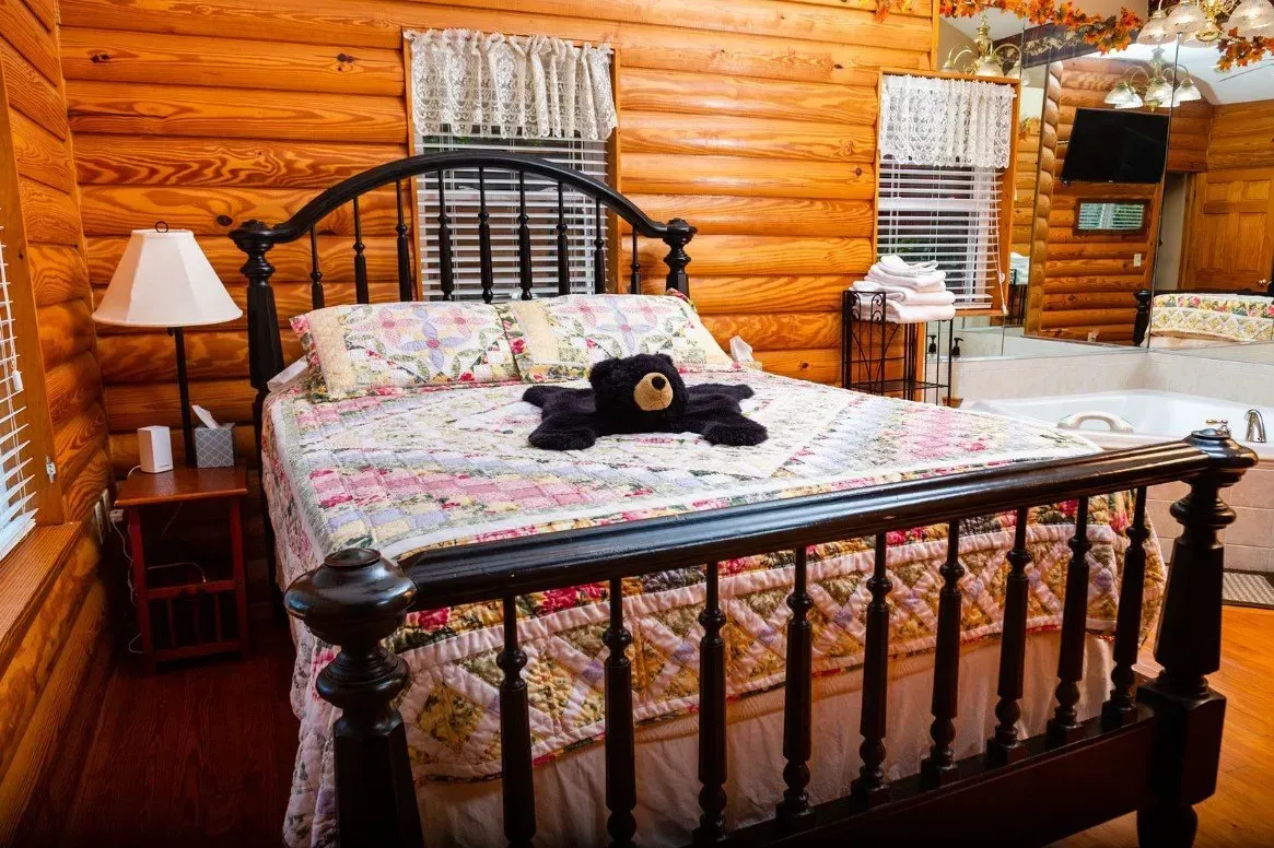 A rustic bedroom with a wooden bed, floral quilt, and a black bear. Log walls and a lamp.
