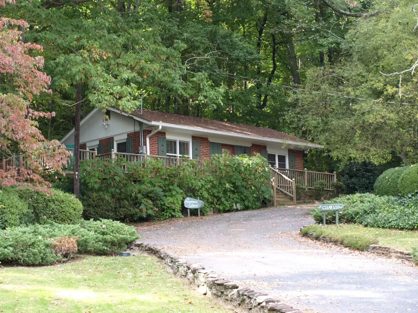 Small brick house with a gravel driveway, surrounded by greenery and trees.