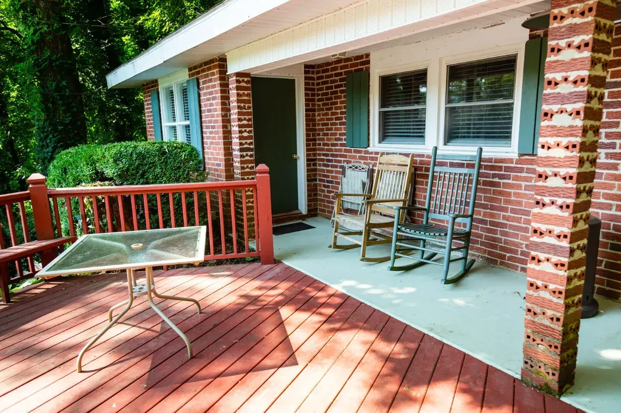 Brick house with a porch, rocking chairs, and a table, surrounded by green foliage.
