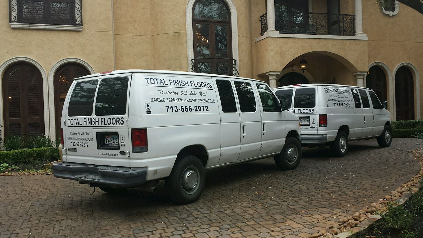 Two white vans parked in front of a large, light-colored house. The vans have company logos.