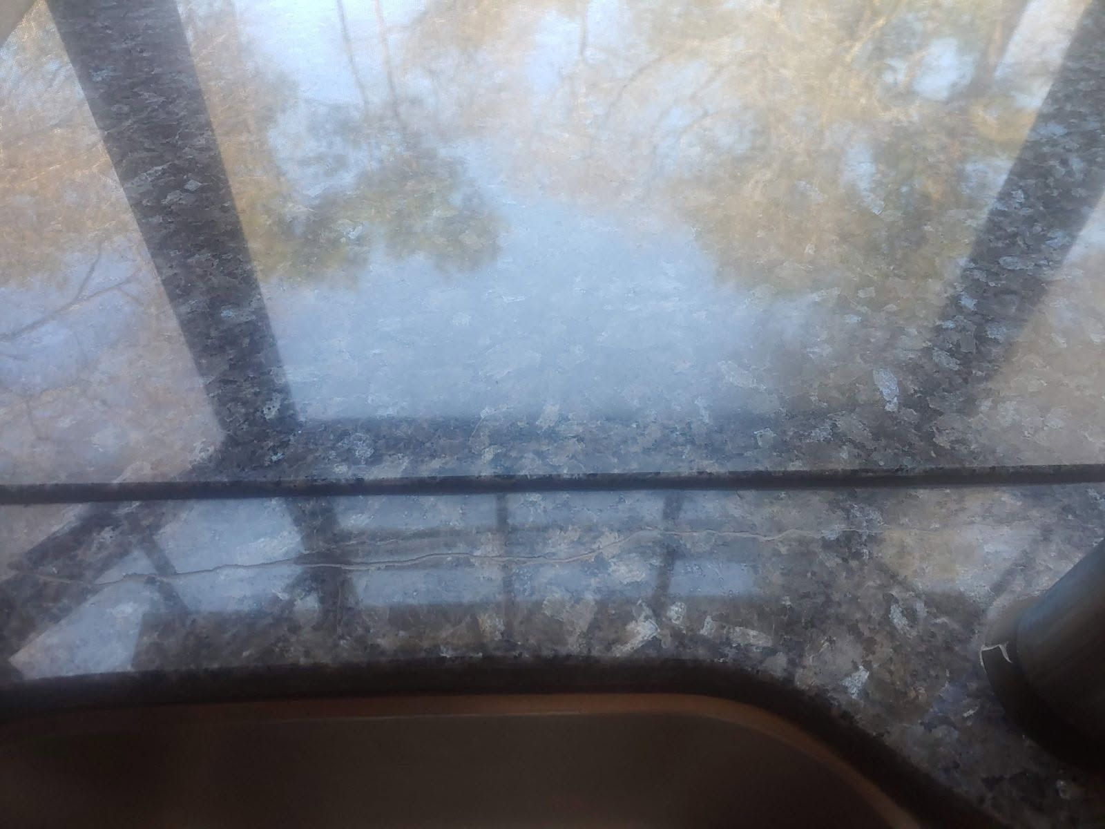 Close-up of a countertop with a sink, reflecting a blurry outdoor scene with trees and light.
