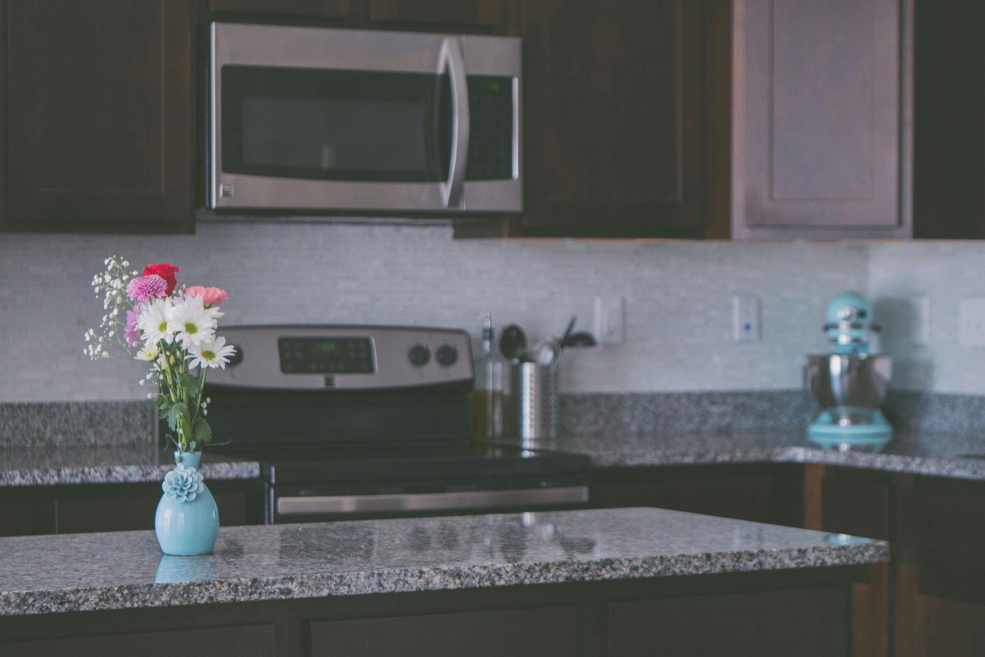 A kitchen scene with flowers, stove, microwave, and stand mixer. Dark cabinets with a granite countertop.
