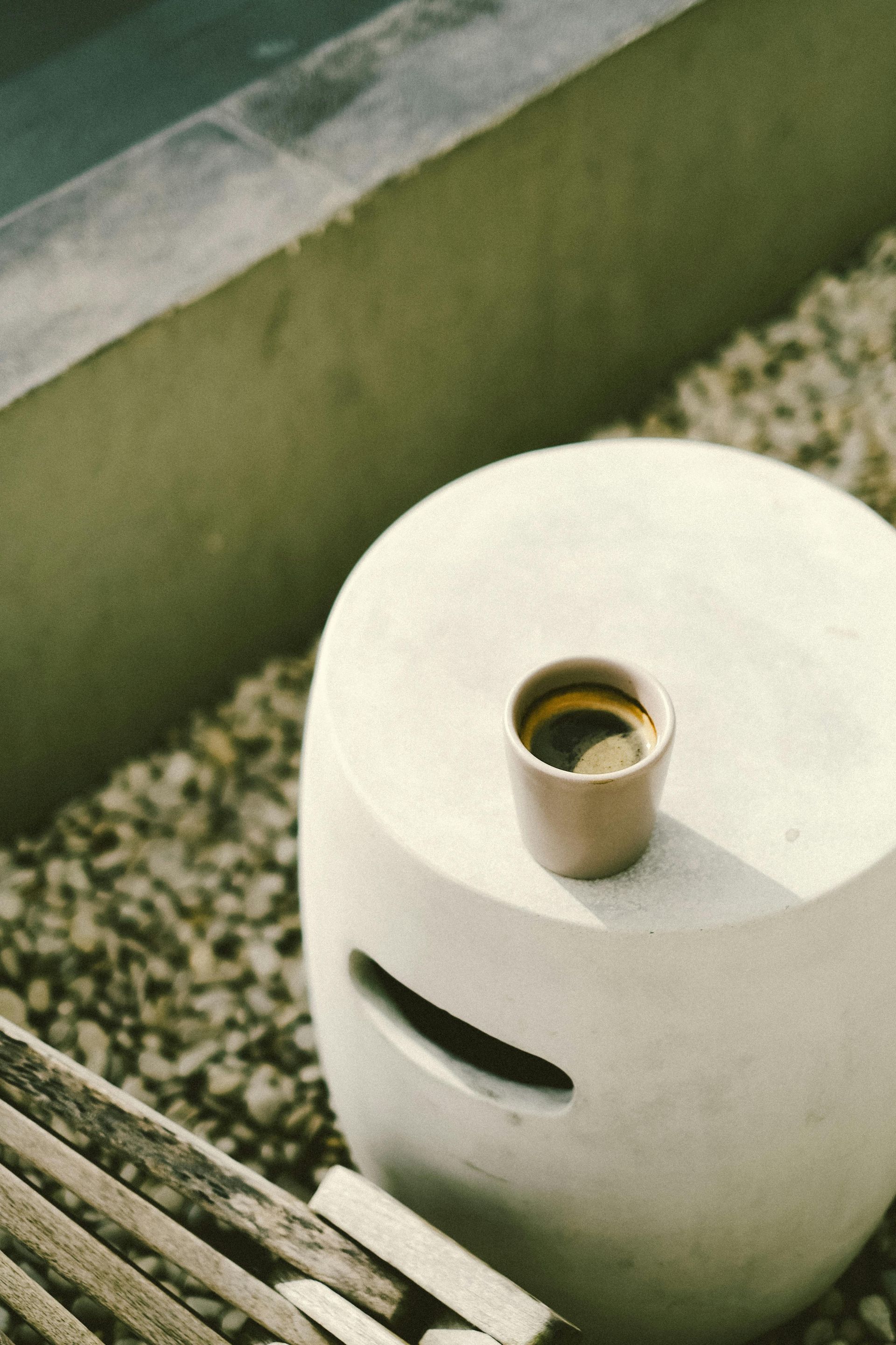 Espresso cup on a white concrete stool, next to pebbles and pool edge.