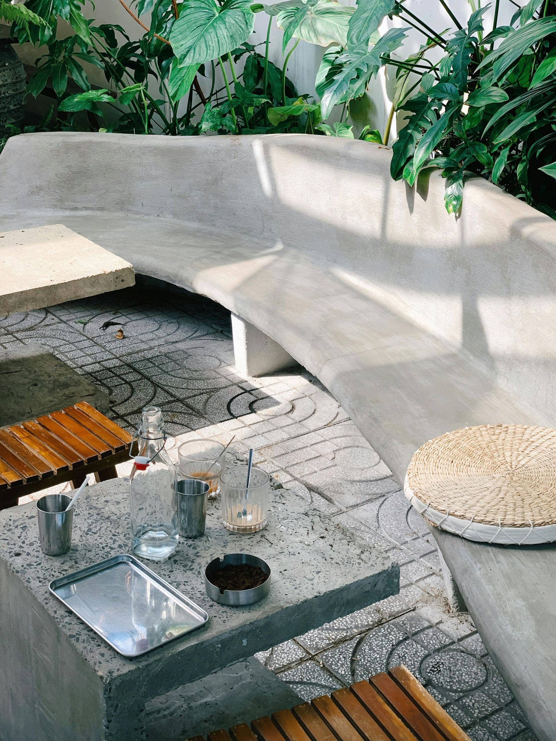 Concrete patio with built-in seating, mosaic tile floor, small wooden tables, and lush green plants.