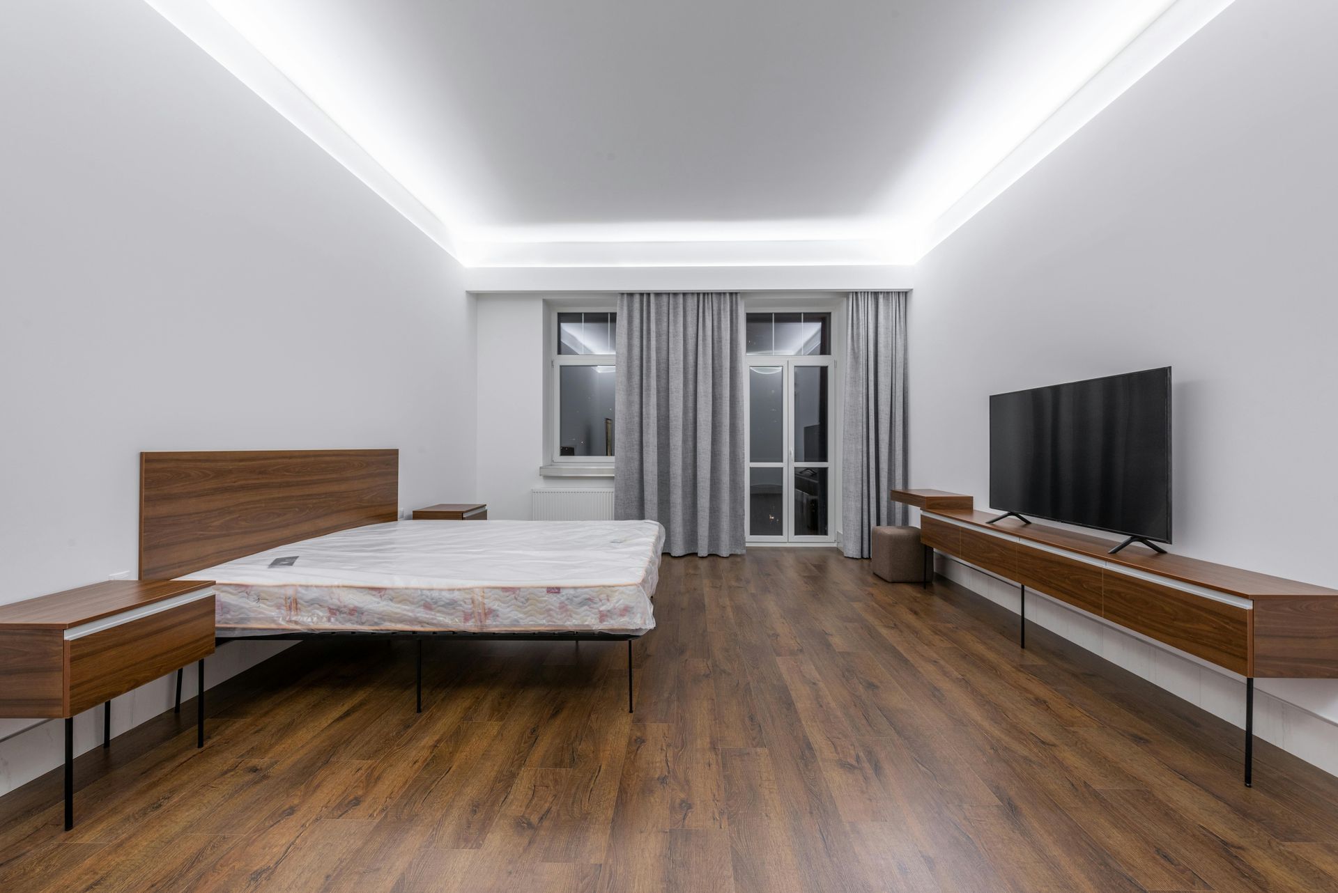 Bedroom with wooden furniture, including bed, nightstands, and TV console, against white walls and dark wood floors.