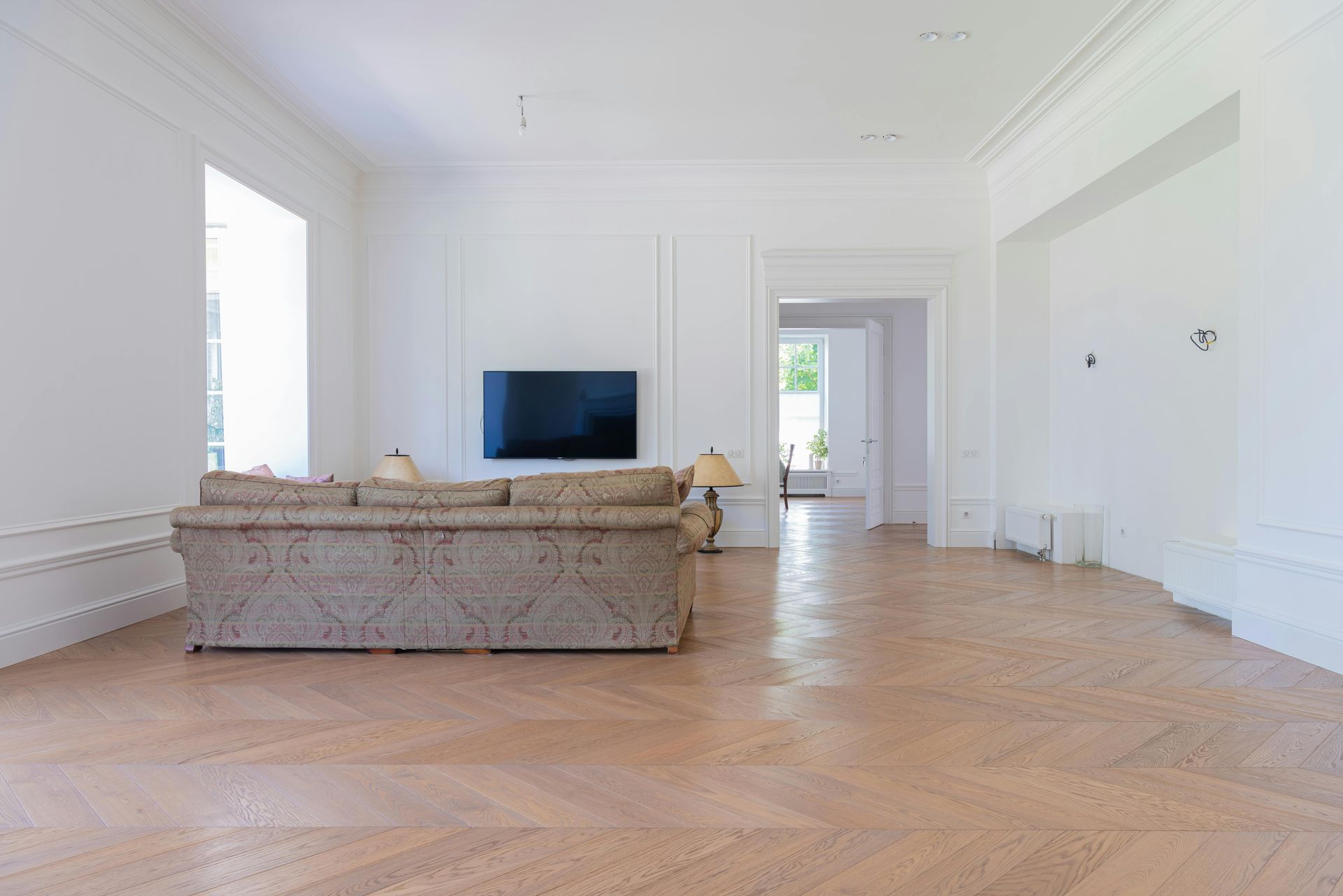 Empty white room with parquet floor, sofa, and mounted TV.