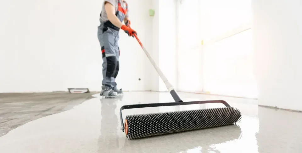 Person using a spiked roller to level epoxy flooring in a room with white walls.