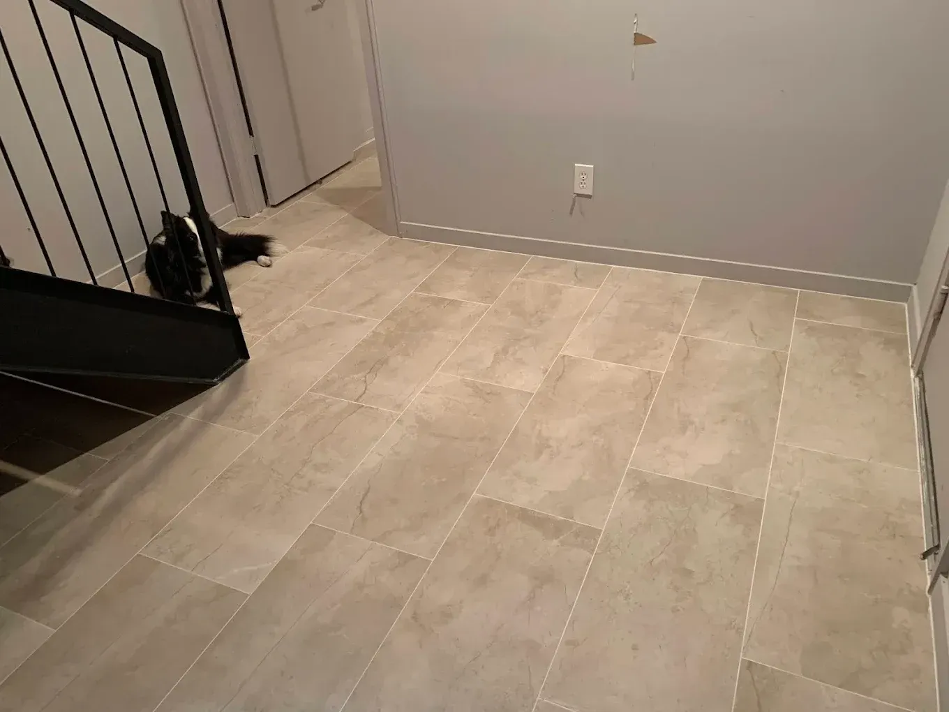 Light tile flooring in a hallway. Stairs with black railing are visible on the left. Gray walls.