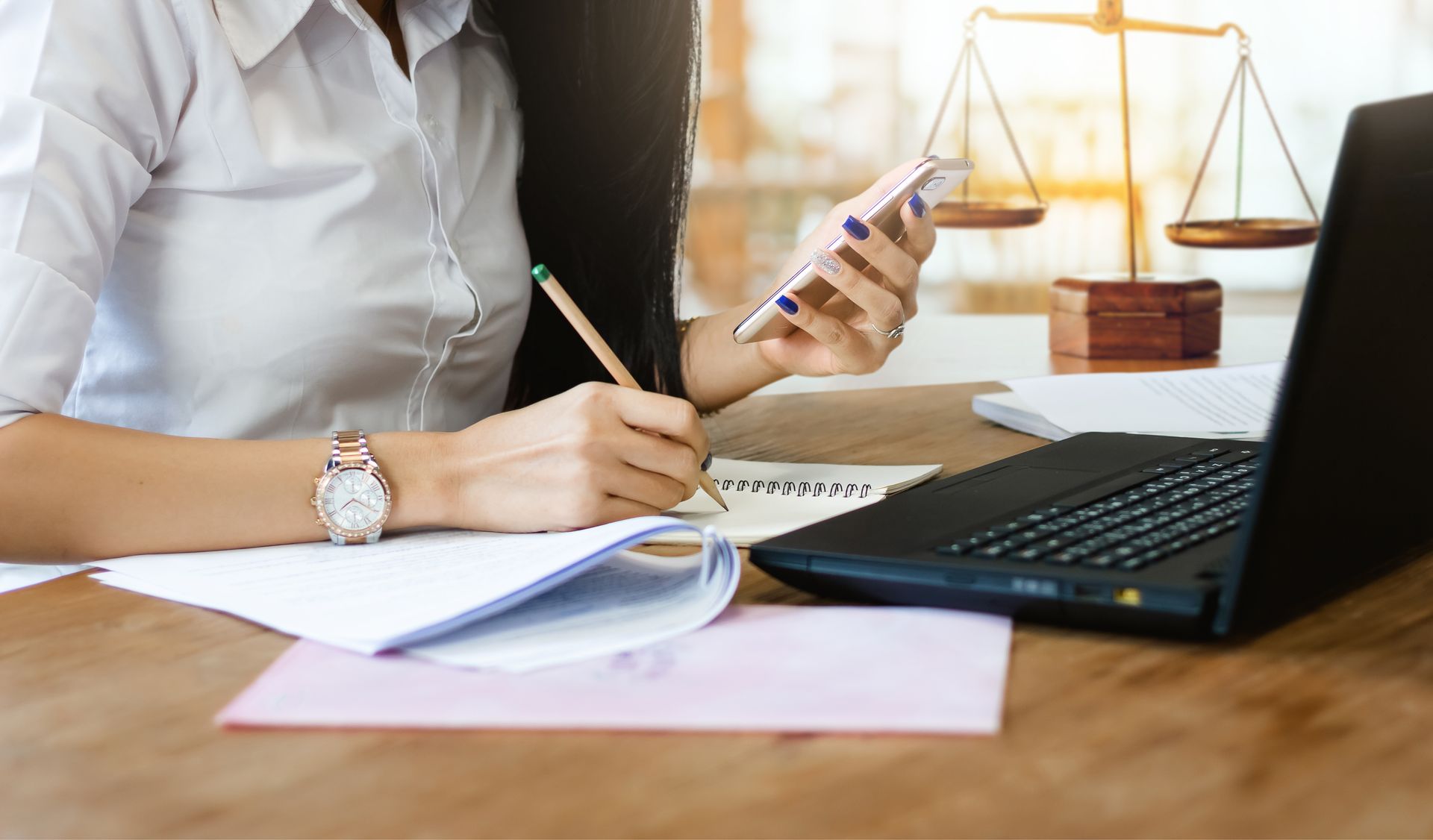 Woman at desk with laptop, phone, and notebook. Scales of justice in background. Woman at desk with laptop, phone, and notebook. Scales of justice in background.